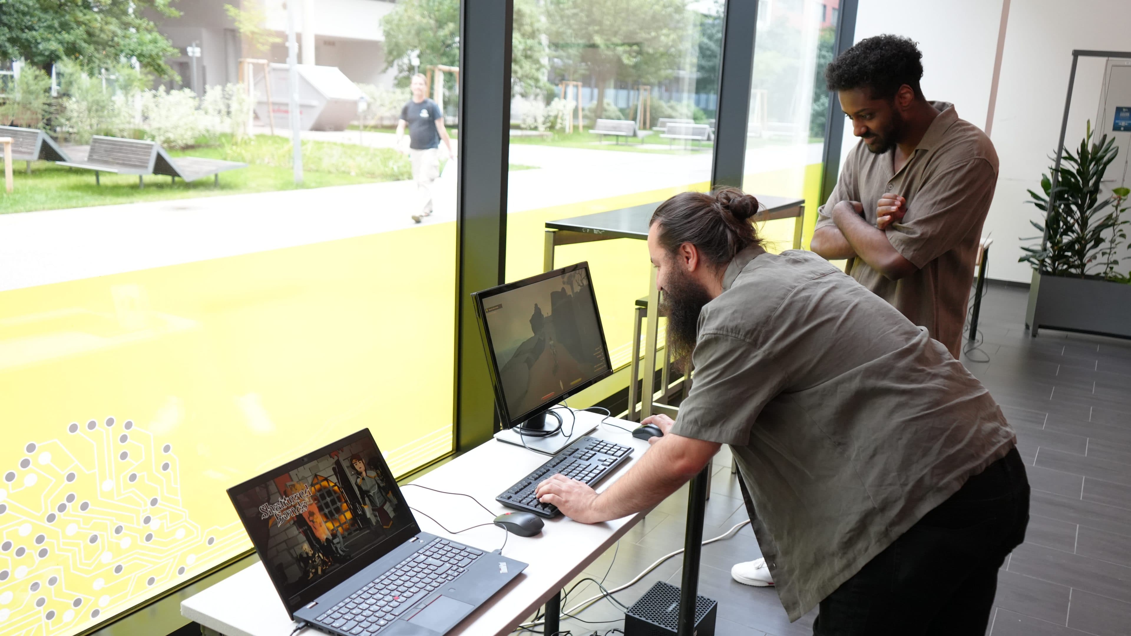 Zwei Männer stehen an einem Tisch neben großen Fenstern und blicken konzentriert auf ihre Computerbildschirme. Der eine tippt auf einer Tastatur, während der andere zusieht. Auf dem Tisch stehen Laptops und ein Desktop-Monitor, und draußen ist Grün zu sehen.