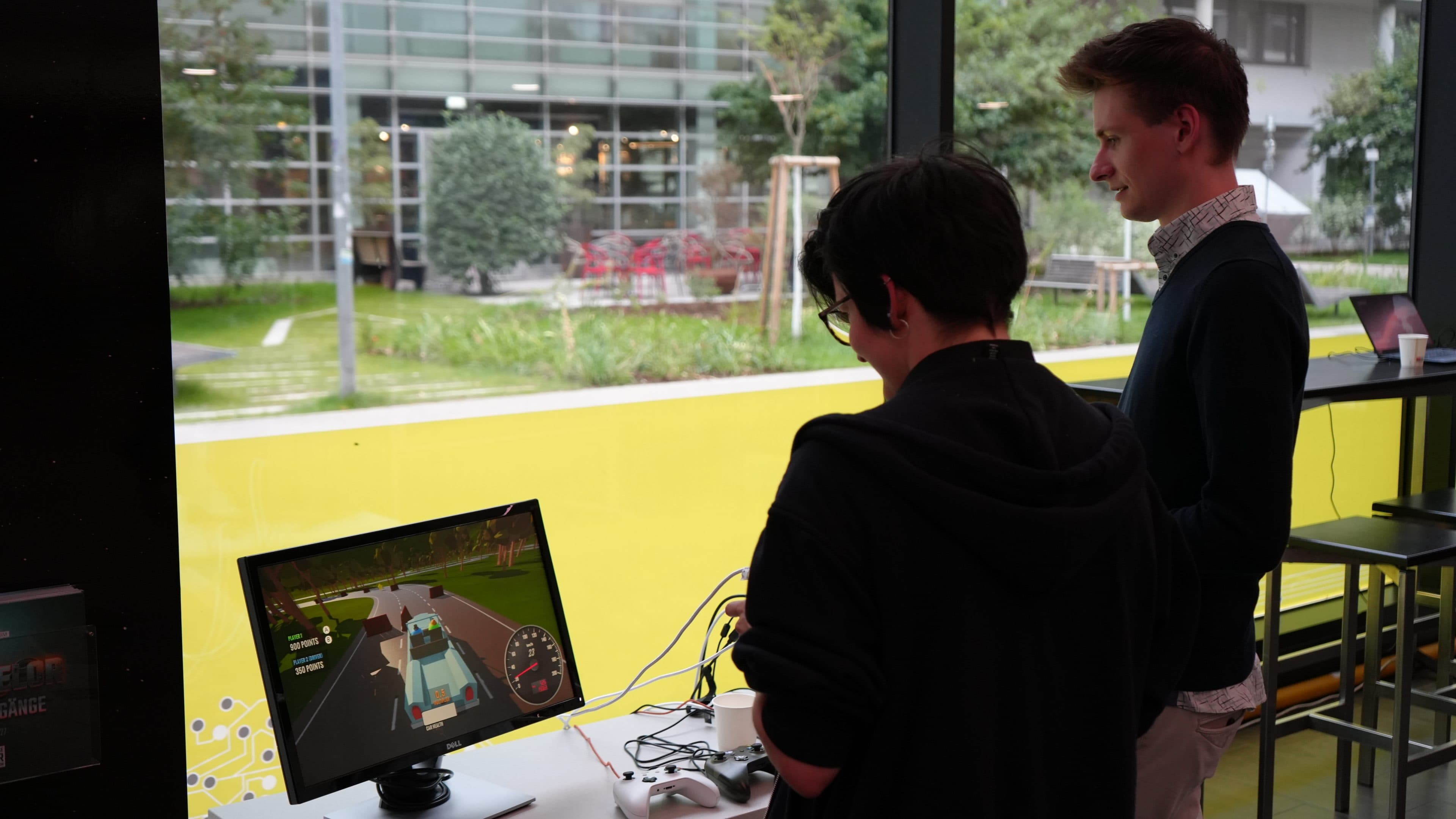 Zwei Personen stehen an einem Tisch neben einem großen Fenster und interagieren mit einer Fahrsimulation, die auf einem Monitor angezeigt wird. Draußen sind Bäume und ein modernes Gebäude zu sehen.