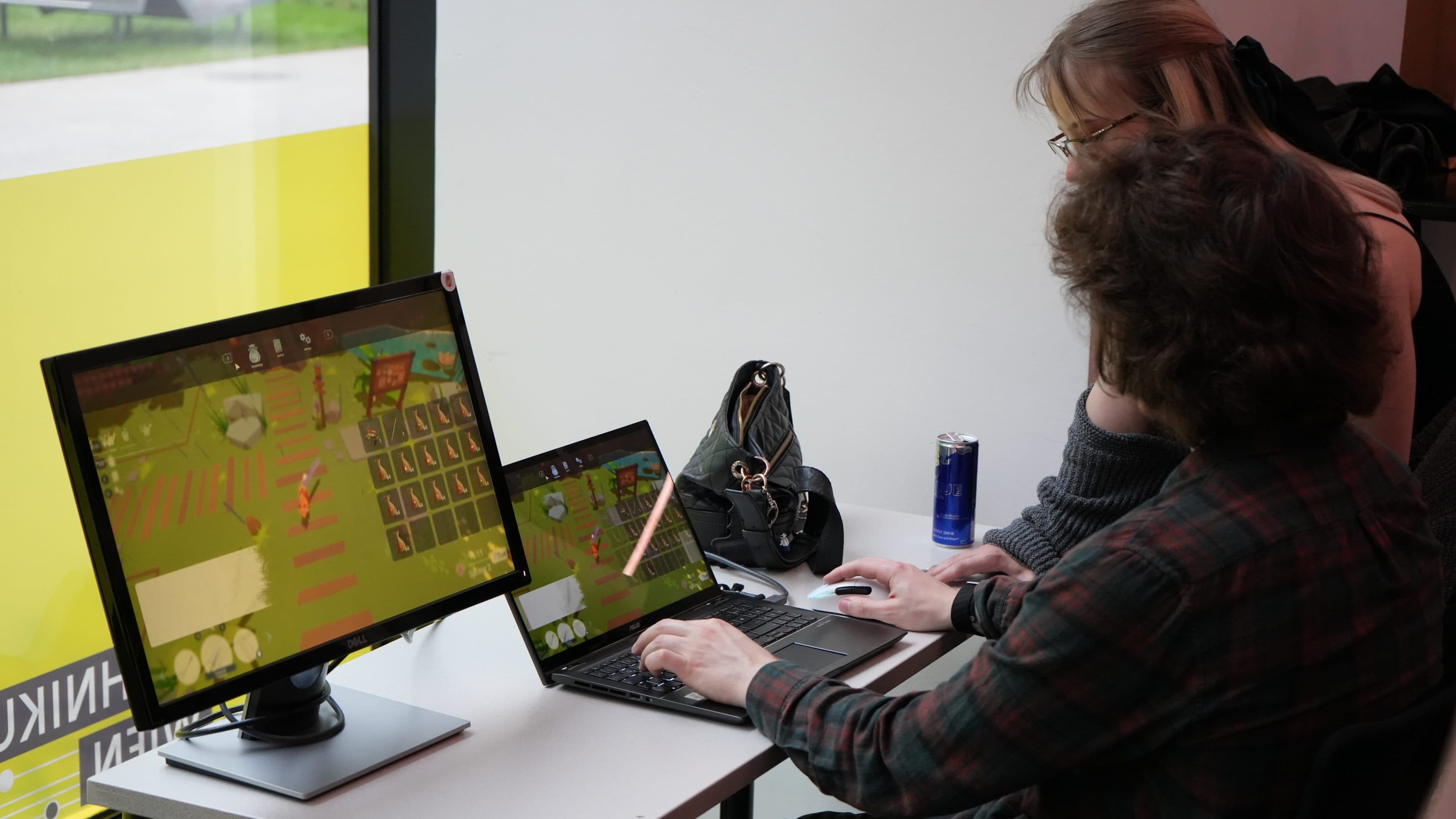 Zwei Personen sitzen an einem Schreibtisch und arbeiten an einem Laptop, der an einen Monitor angeschlossen ist; auf beiden Bildschirmen ist dasselbe farbenfrohe Videospiel zu sehen. Auf dem Schreibtisch neben ihnen, direkt neben einem großen Fenster, liegen eine Handtasche und eine Dose.