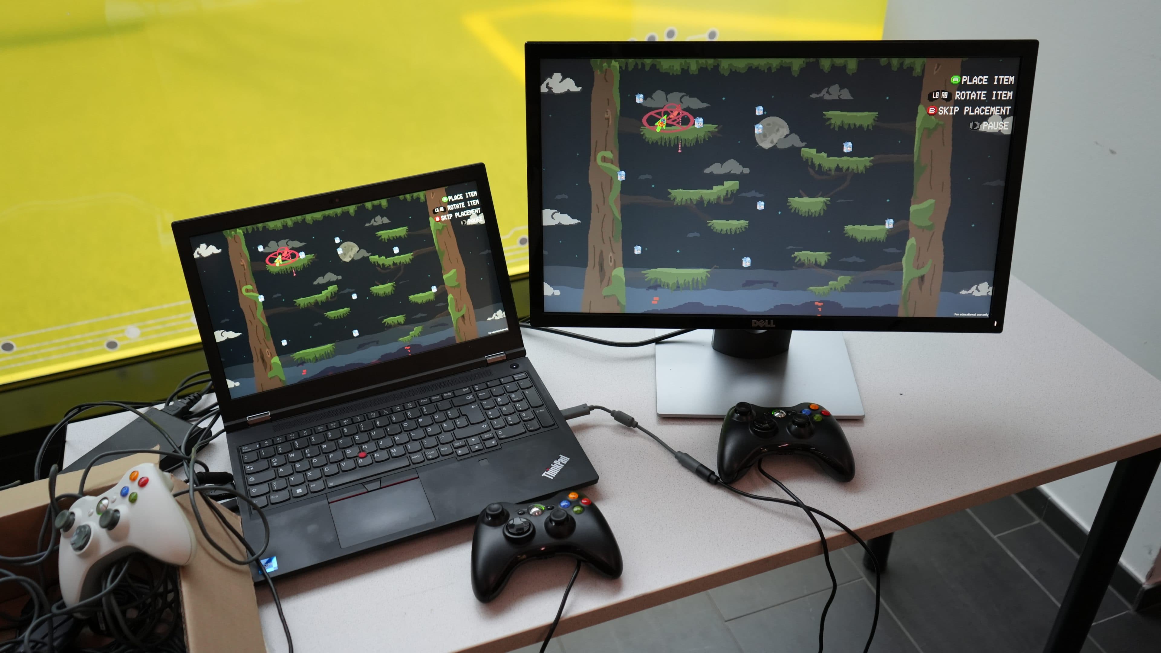 Auf einem Laptop und einem externen Monitor ist dieselbe Pixel-Art-Szene aus einem Videospiel zu sehen. Zwei Xbox-Controller und ein PlayStation-Controller sind angeschlossen und liegen auf dem Schreibtisch. Im Hintergrund ist ein gelbes Fenster zu sehen.