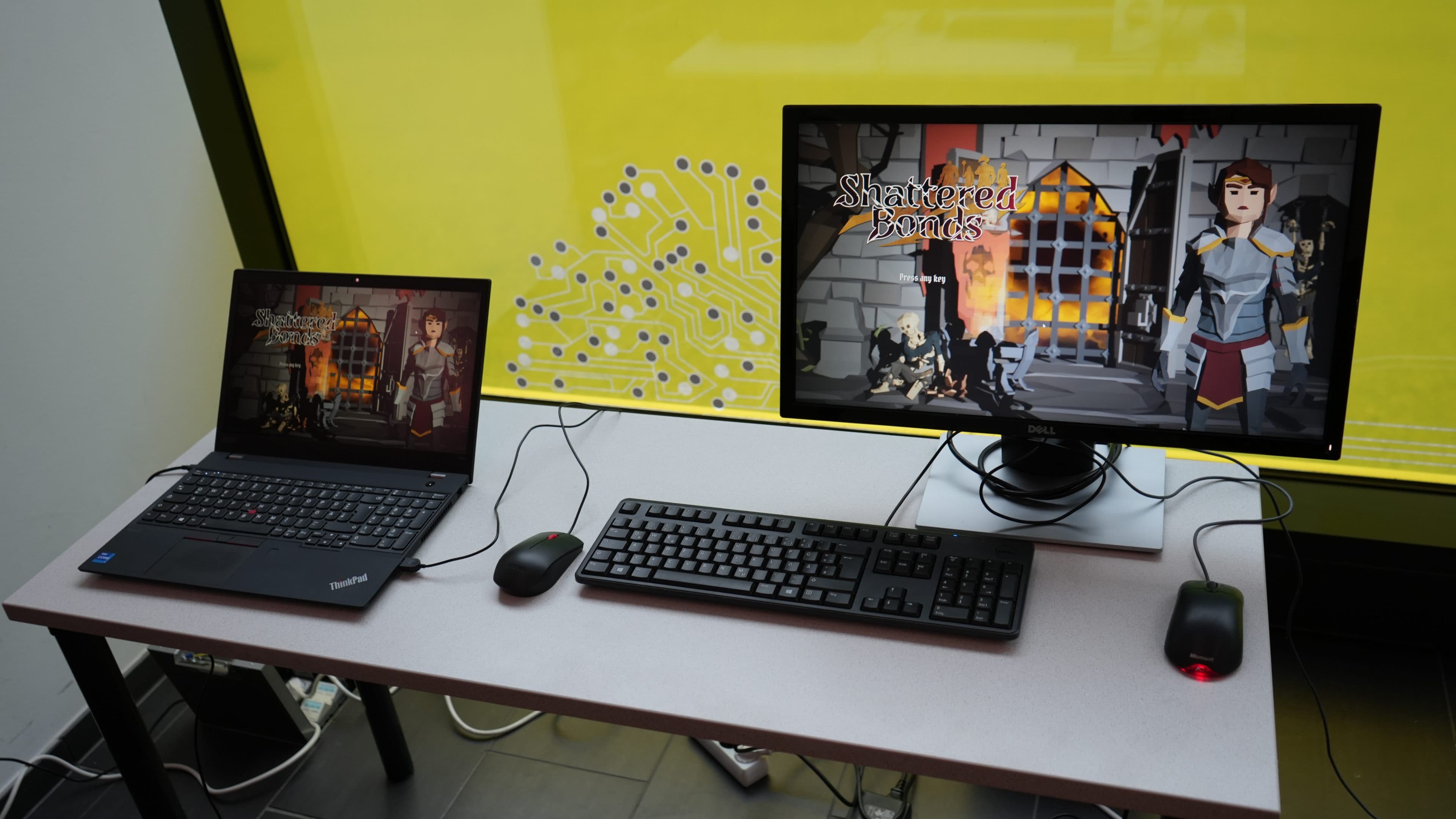 Auf einem Tisch, vor dem eine Tastatur und zwei Mäuse stehen, sind ein Laptop und ein Desktop-Monitor zu sehen, auf denen dasselbe mittelalterlich angehauchte Spiel, „Shattered Bonds“, läuft; der Hintergrund ist gelb und mit einer Schaltkreisgrafik versehen.