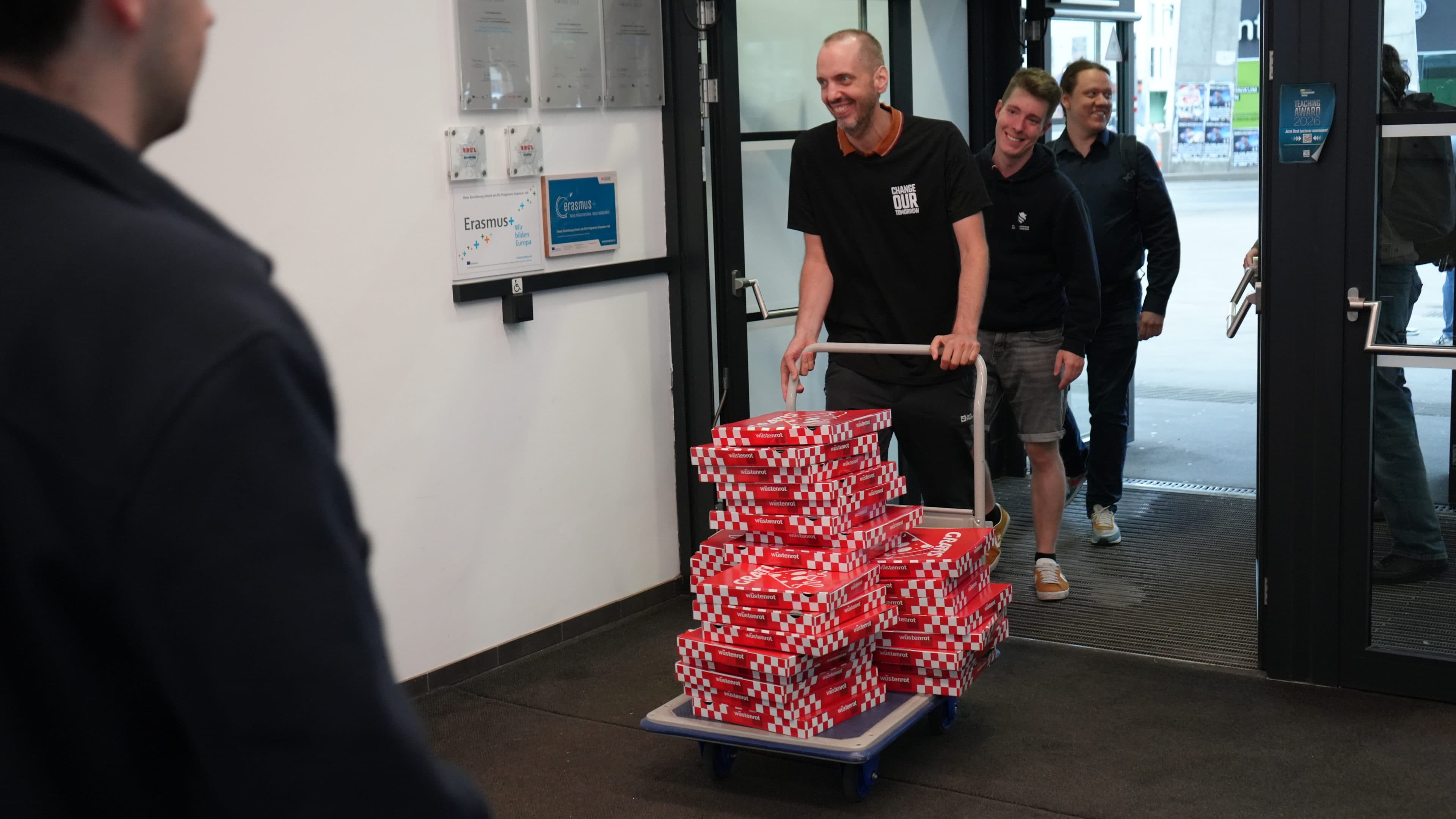 Ein Mann schiebt einen mit Pizzakartons beladenen Wagen durch eine Tür, gefolgt von drei lächelnden Personen. Im Vordergrund steht eine weitere Person, die nur teilweise zu sehen ist.