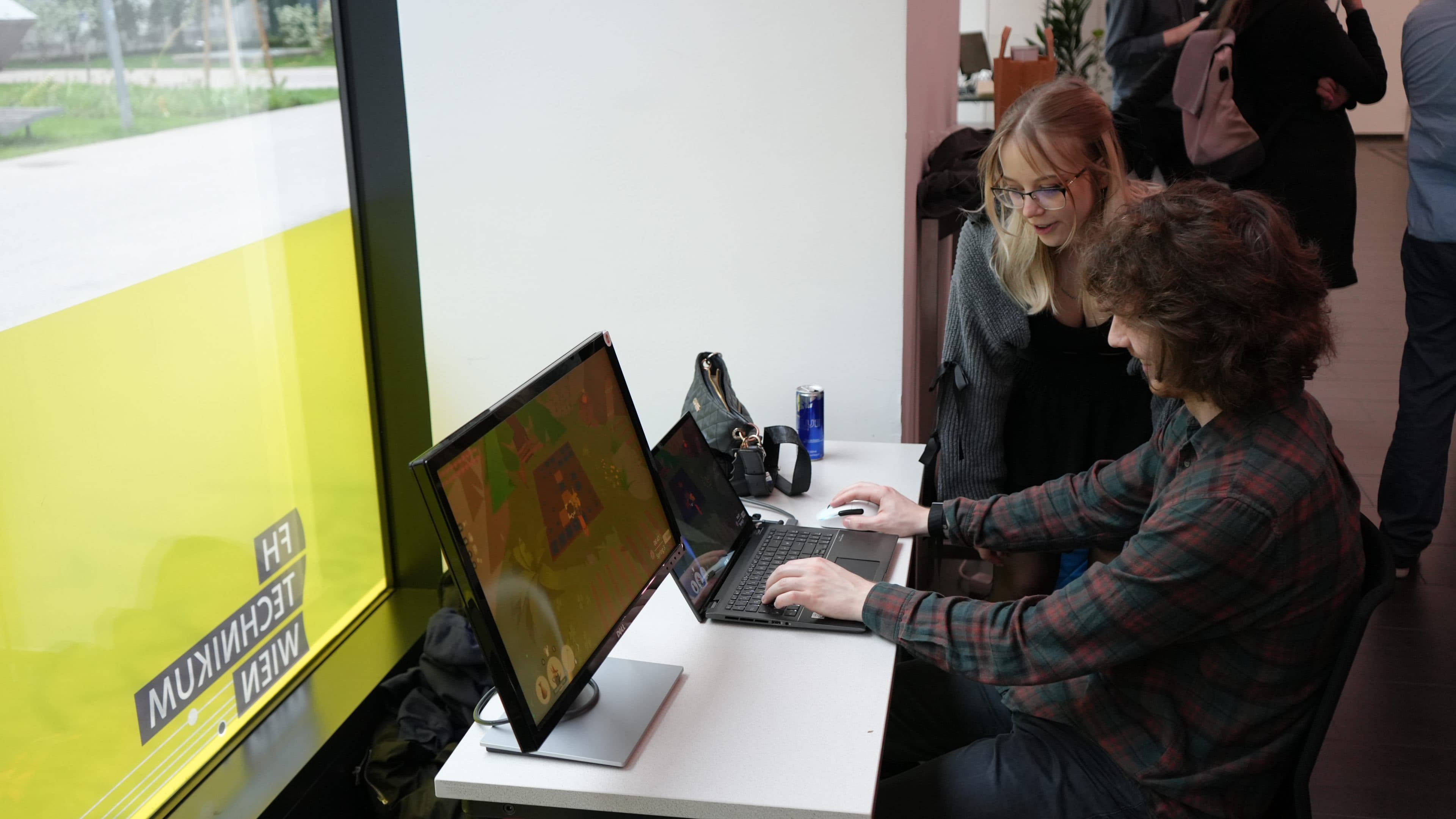 Ein Mann und eine Frau sitzen an einem Schreibtisch, lächeln und schauen gemeinsam auf einen Computerbildschirm. Der Mann tippt auf einer Tastatur, und auf dem Monitor ist ein Spiel zu sehen. Links ist ein großes gelbes Fenster zu sehen.