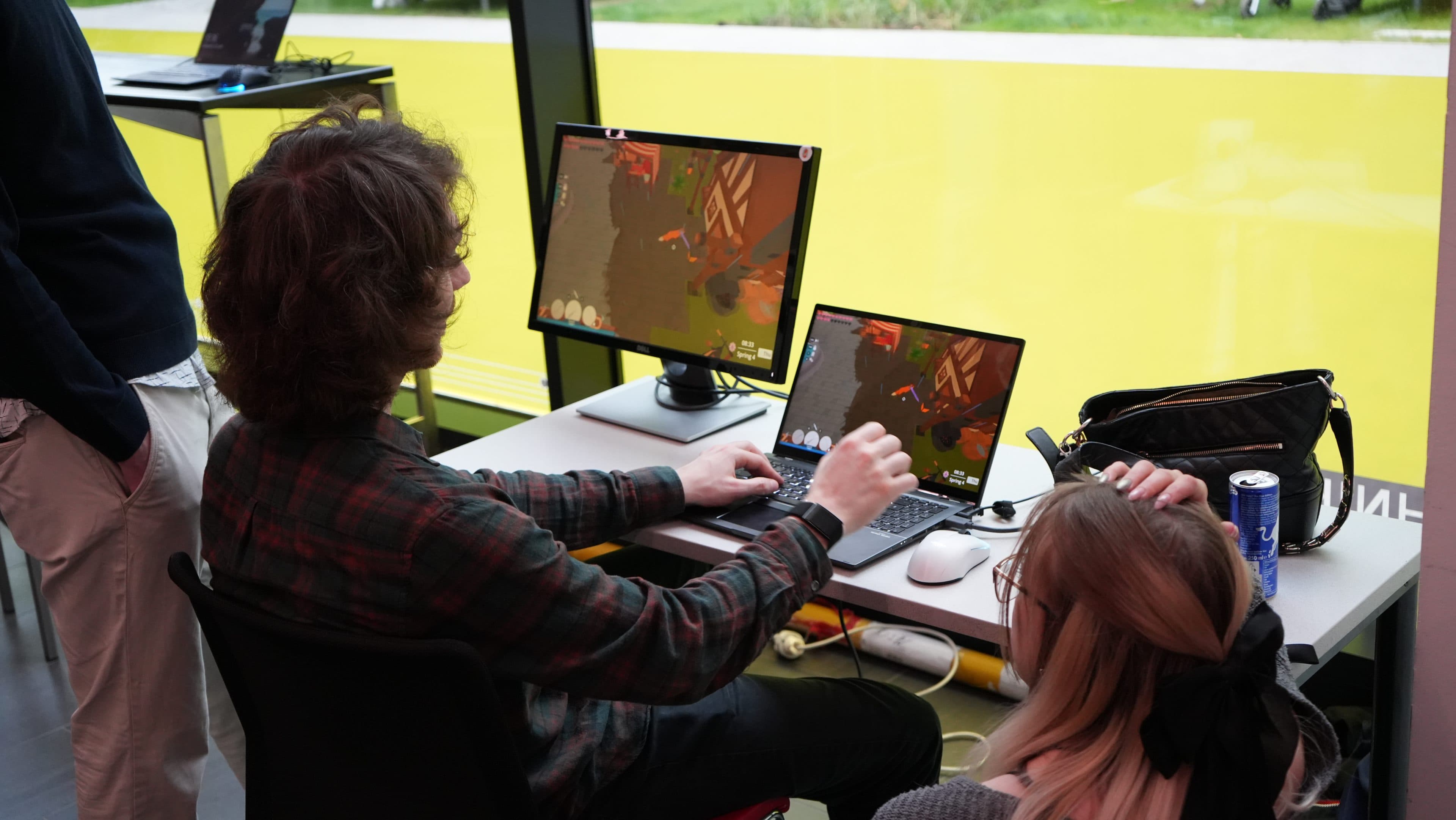 Zwei Personen sitzen an einem Schreibtisch und spielen auf zwei Monitoren ein Videospiel. Eine Person bedient Tastatur und Maus, während die andere zusieht. Auf dem Schreibtisch liegen eine Tasche, Kopfhörer und eine Dose Red Bull. Im Hintergrund ist eine leuchtend gelbe Wand zu sehen.