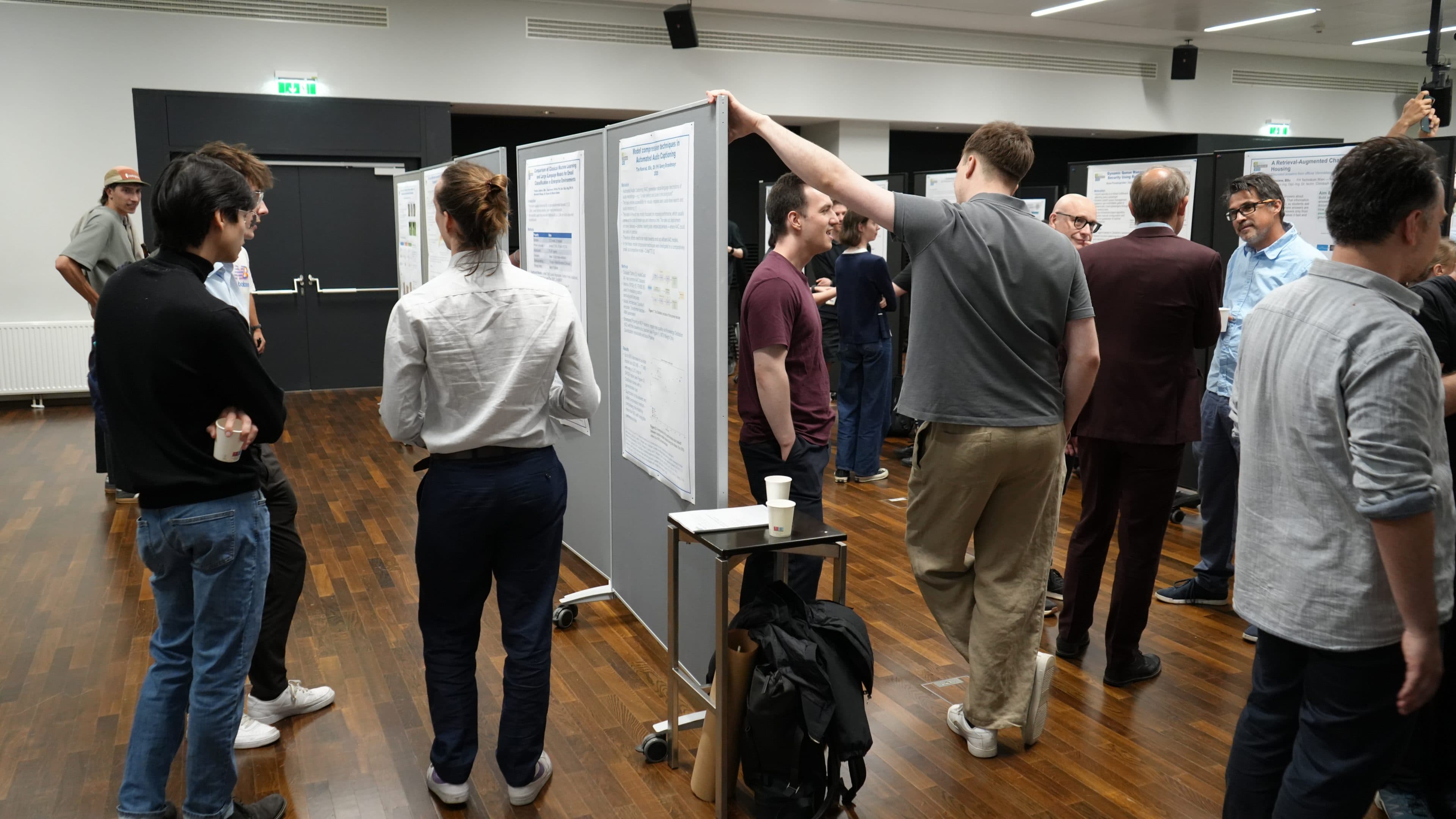 Eine Gruppe von Menschen steht in einem Konferenzraum und diskutiert über Forschungsplakate, die an Tafeln ausgestellt sind; einige unterhalten sich zu zweit, während andere die Plakate lesen. Die Atmosphäre ist ungezwungen und von Zusammenarbeit geprägt.