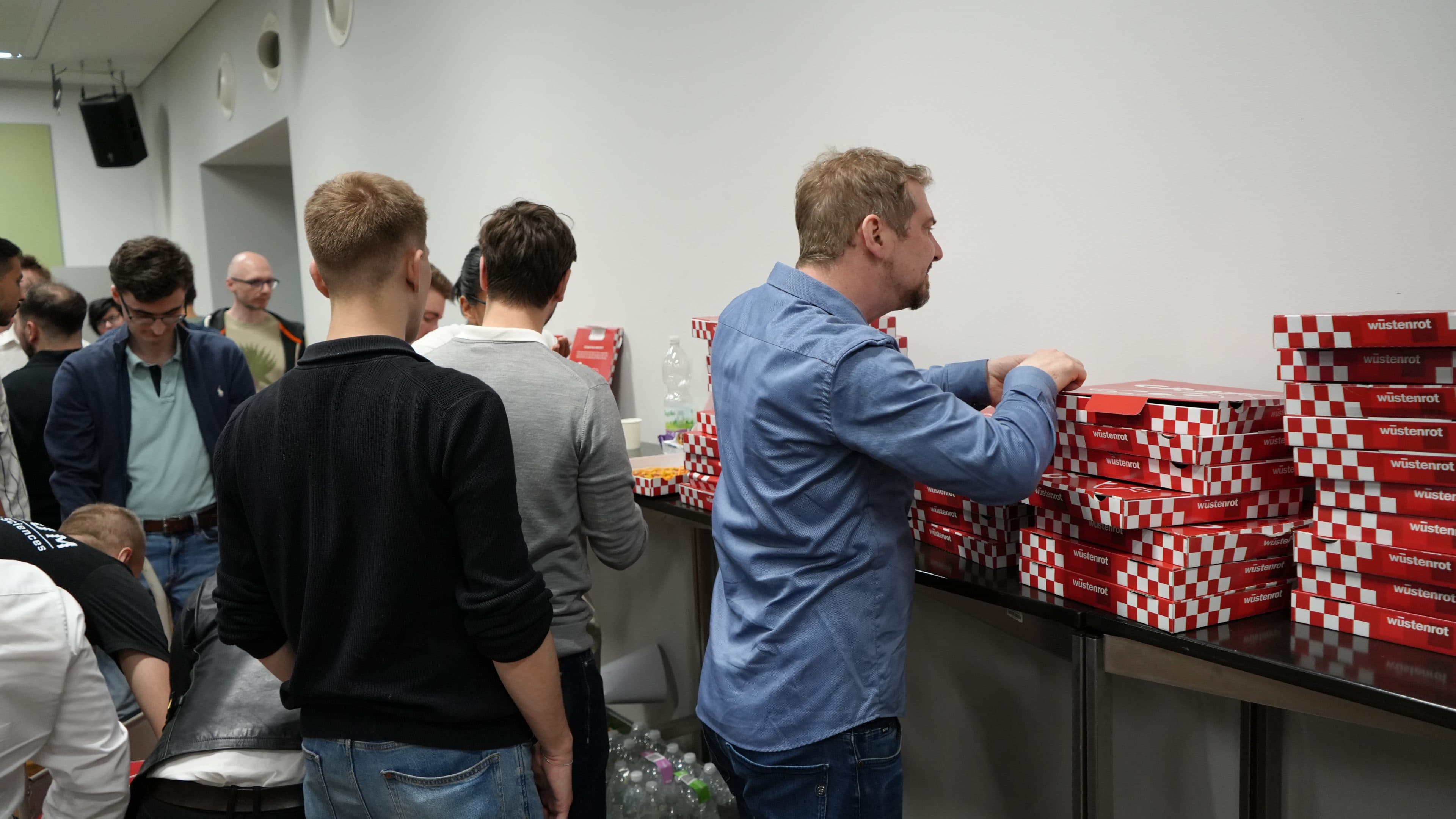 Mehrere Leute stehen in einer Schlange neben einer Theke, auf der rot-weiße Pizzakartons gestapelt sind. Ein Mann öffnet gerade einen Karton, während andere hinter ihm warten. Unter der Theke sind Flaschen und Getränke zu sehen.