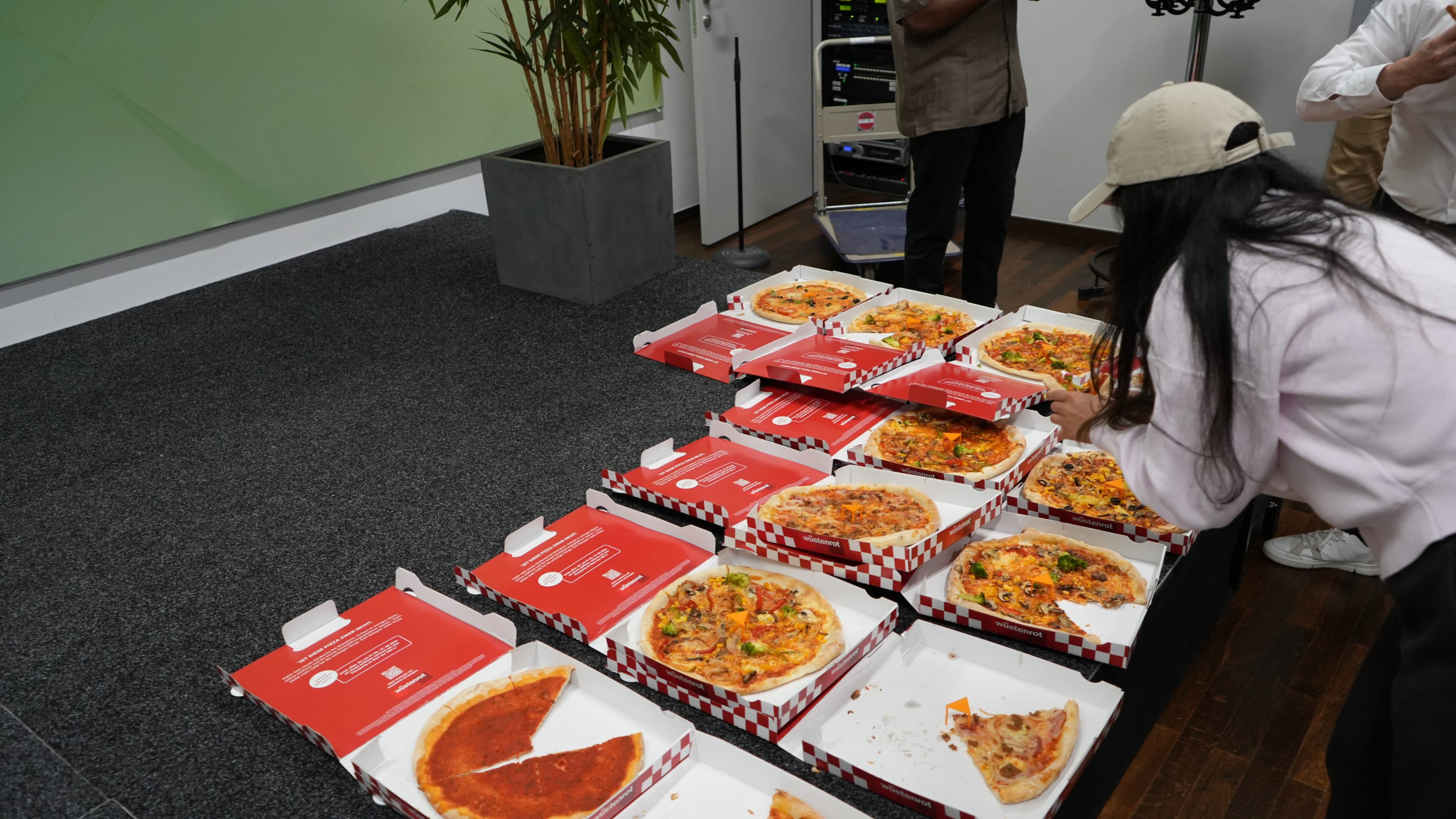 Auf einem Tisch stehen mehrere offene Pizzakartons mit verschiedenen Pizzasorten. Eine Frau mit weißer Mütze und rosa Shirt nimmt sich ein Stück, während andere Leute daneben stehen. Im Hintergrund ist eine Pflanze zu sehen.