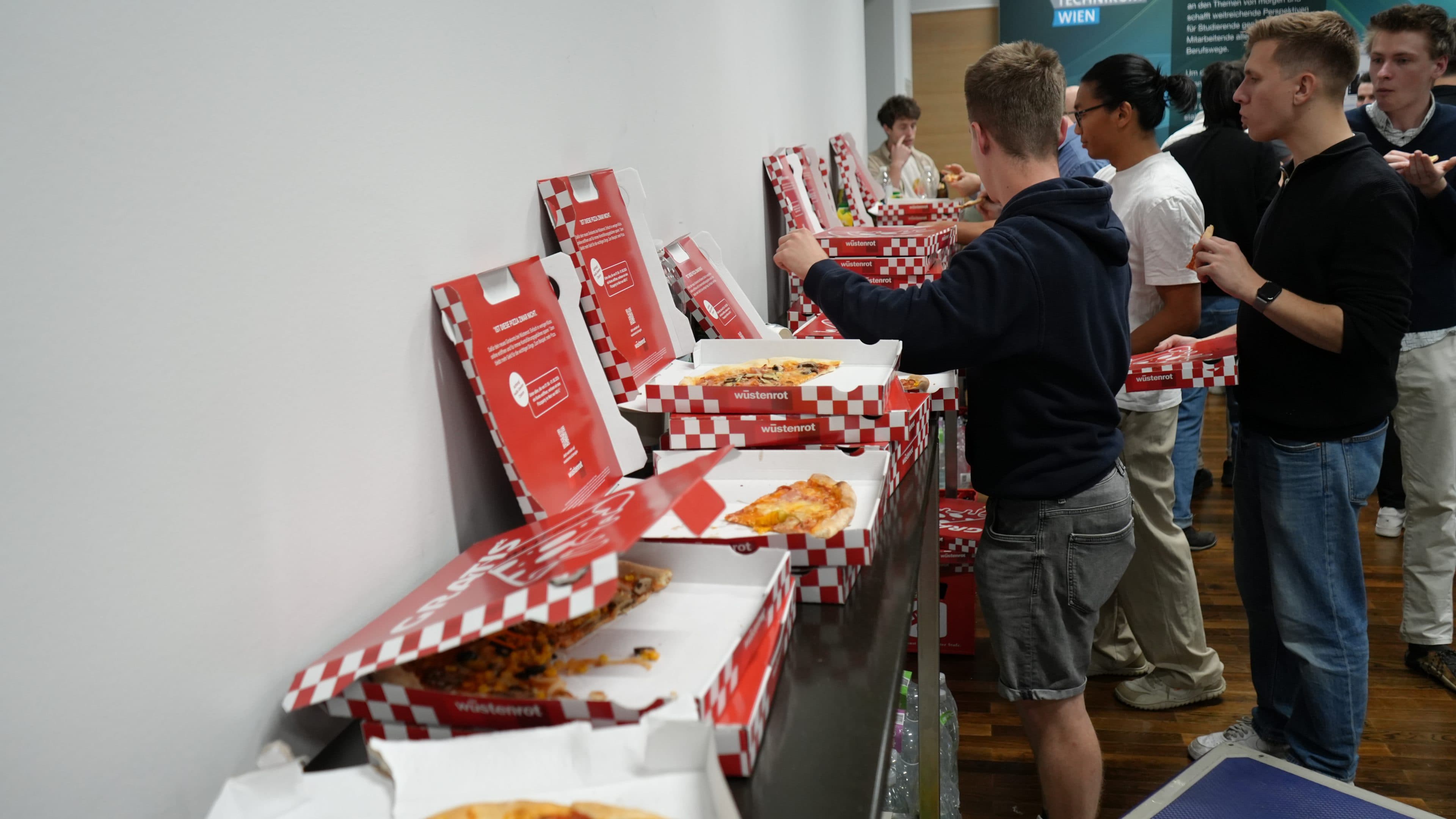 Mehrere Personen stehen an einem langen Tisch, auf dem offene Pizzakartons mit rot-weiß karierten Deckeln stehen, und nehmen sich ein Stück. Die Szene wirkt wie eine ungezwungene Veranstaltung oder ein Treffen in einem Innenraum.