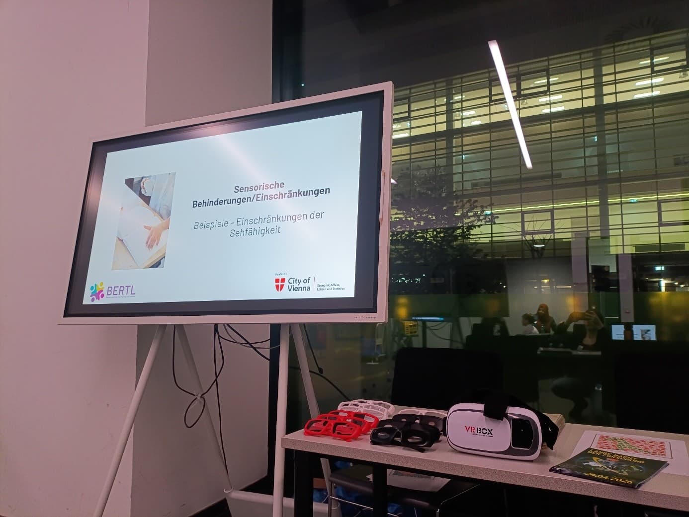 Auf einem Monitor wird eine Präsentationsfolie auf Deutsch zum Thema Sinnesbeeinträchtigungen angezeigt. Daneben liegen auf einem Tisch Hilfsmittel, darunter ein VR-Headset und eine Brille. Im Hintergrund sitzen Menschen nachts an großen Fenstern.
