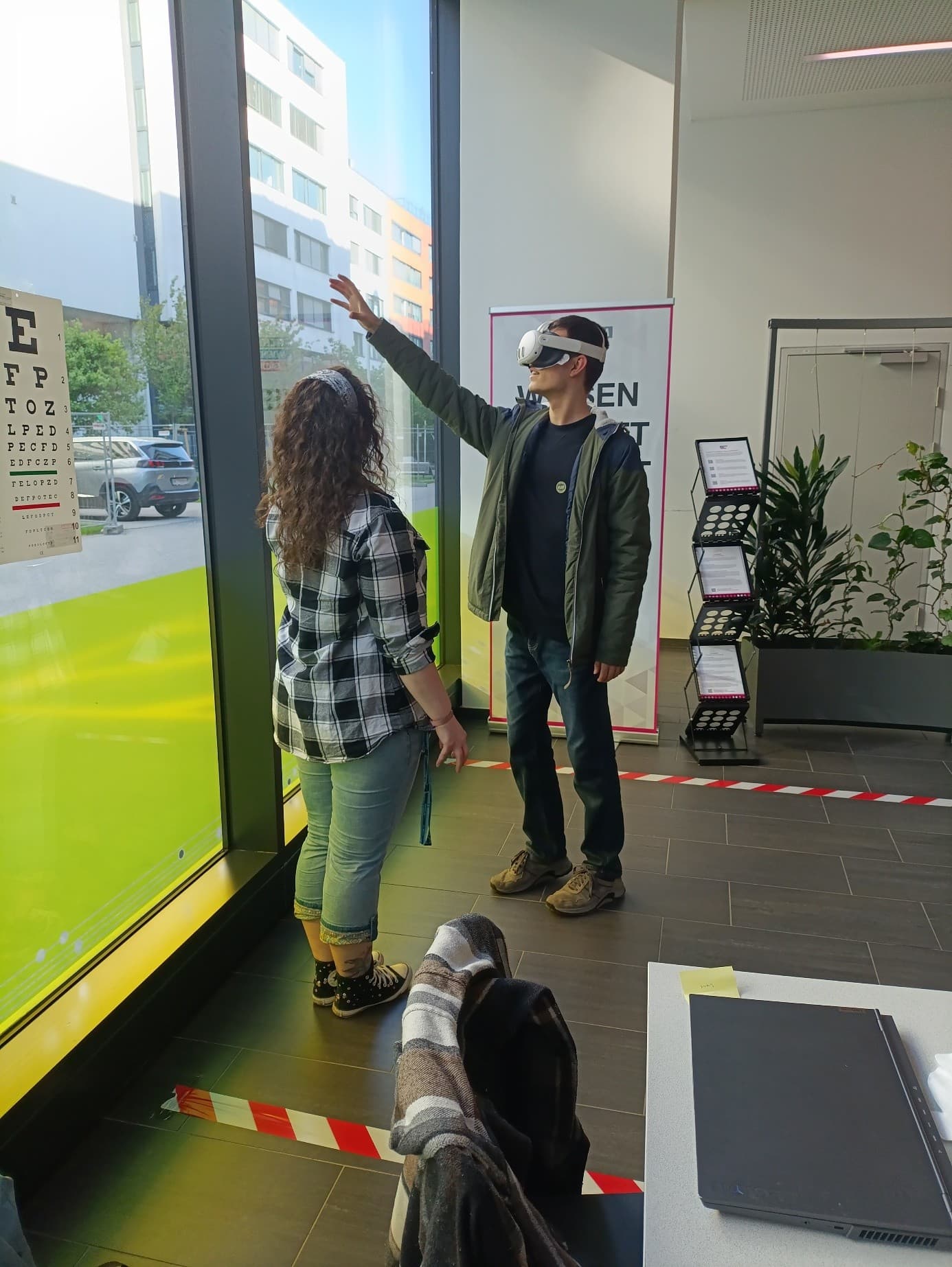 Ein Mann mit einer Virtual-Reality-Brille hebt den Arm, während eine Frau neben ihm in einem modernen Raum mit großen Fenstern steht. Beide befinden sich in der Nähe von roten Klebebandmarkierungen auf dem Boden, und im Hintergrund sind Büromöbel und Pflanzen zu sehen.