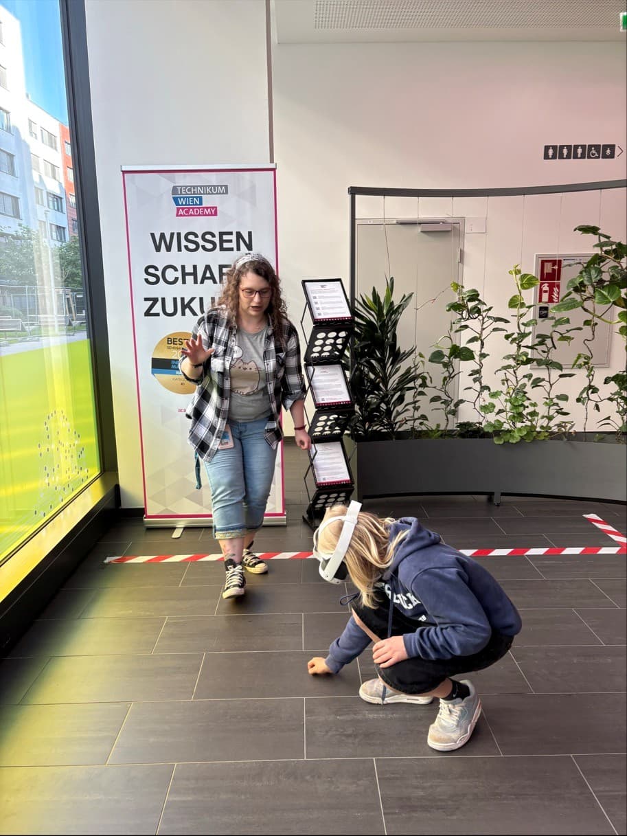 Zwei junge Menschen befinden sich in einem modernen Gebäude; einer steht lächelnd da und macht ein Peace-Zeichen, während der andere neben einem rot-weißen Absperrband auf dem Boden hockt. Im Hintergrund sind ein Schild und Pflanzen zu sehen.