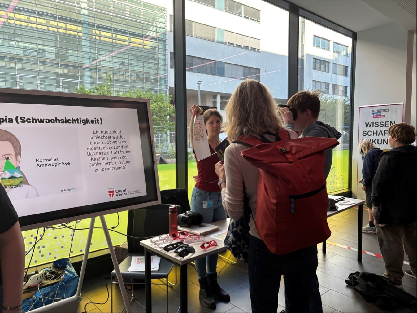 Bei einer Wissenschaftsveranstaltung stehen Menschen in einem modernen Gebäude. Eine Frau mit einem roten Rucksack steht vor einem Informationsstand, an dem ein Plakat zum Thema Amblyopie ausgestellt ist, auf dem Bilder zu sehen sind, die ein normales Auge mit einem amblyopen Auge vergleichen.