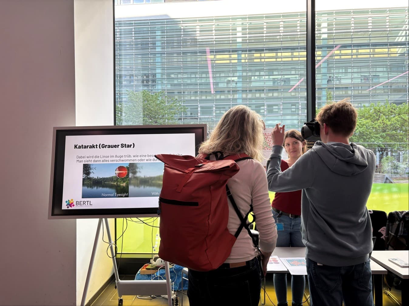 Drei Personen unterhalten sich an einem Stand in einem Innenraum, an dem auf einem großen Bildschirm Informationen zum Thema Grauer Star angezeigt werden. Eine Frau mit einem roten Rucksack steht mit dem Gesicht zum Bildschirm, während ein junger Mann neben ihr gestikuliert. Durch eine Glaswand ist draußen ein Bürogebäude zu sehen.