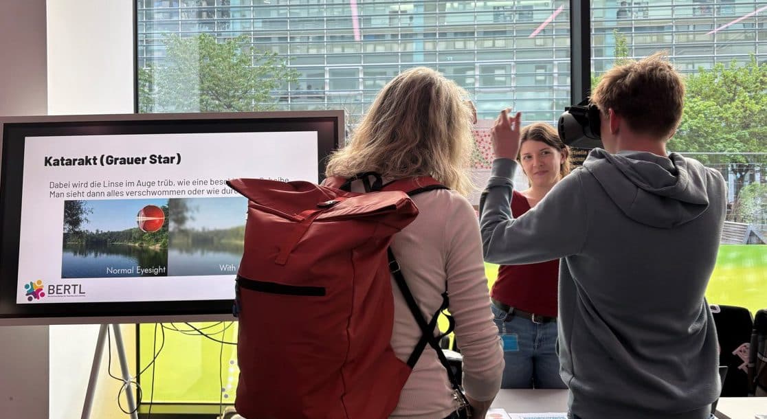 Drei Personen unterhalten sich an einem Stand in einem Innenraum. Auf einem Bildschirm werden Informationen zum Thema Grauer Star auf Deutsch angezeigt. Durch die großen Fenster hinter ihnen sind ein modernes Gebäude und Bäume im Freien zu sehen.