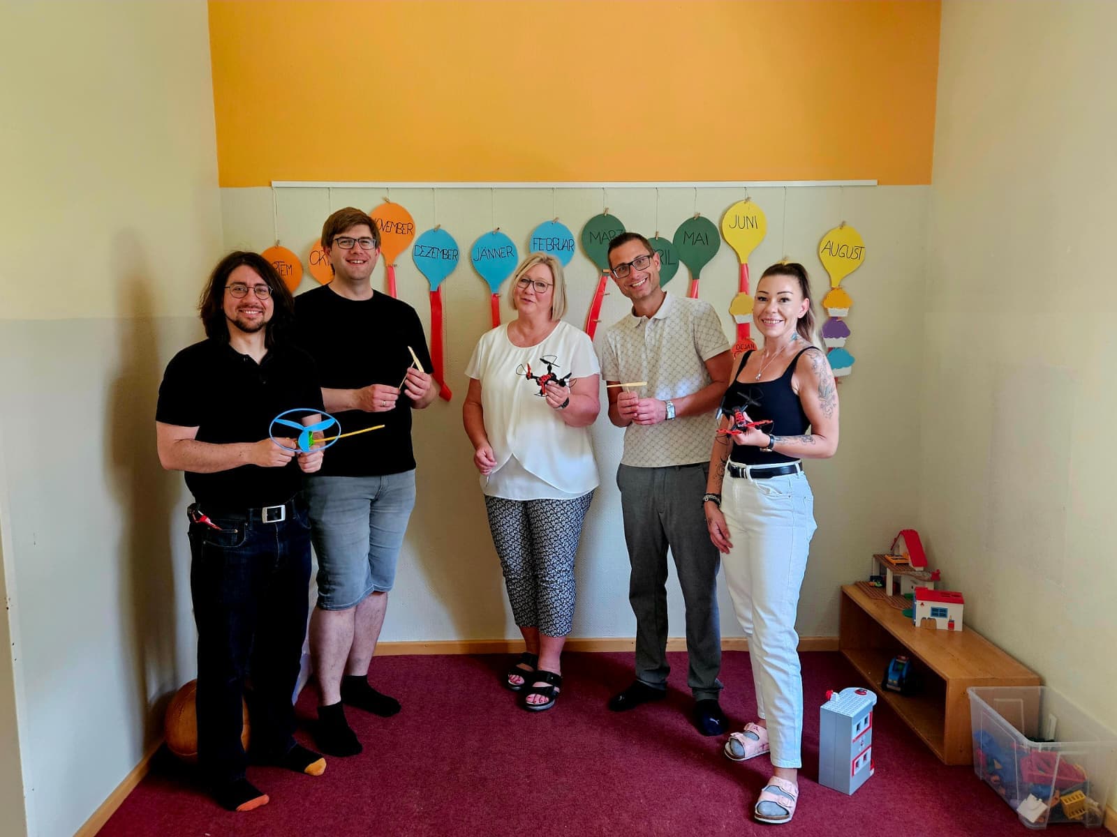 Fünf Erwachsene stehen lächelnd in einem farbenfrohen Raum, hinter ihnen hängen an der Wand Luftballons, die mit den Monaten beschriftet sind. Sie halten kleine Gegenstände oder Bastelarbeiten in den Händen und wirken entspannt und fröhlich. In der Ecke stehen Spielsachen und ein Puppenhaus.