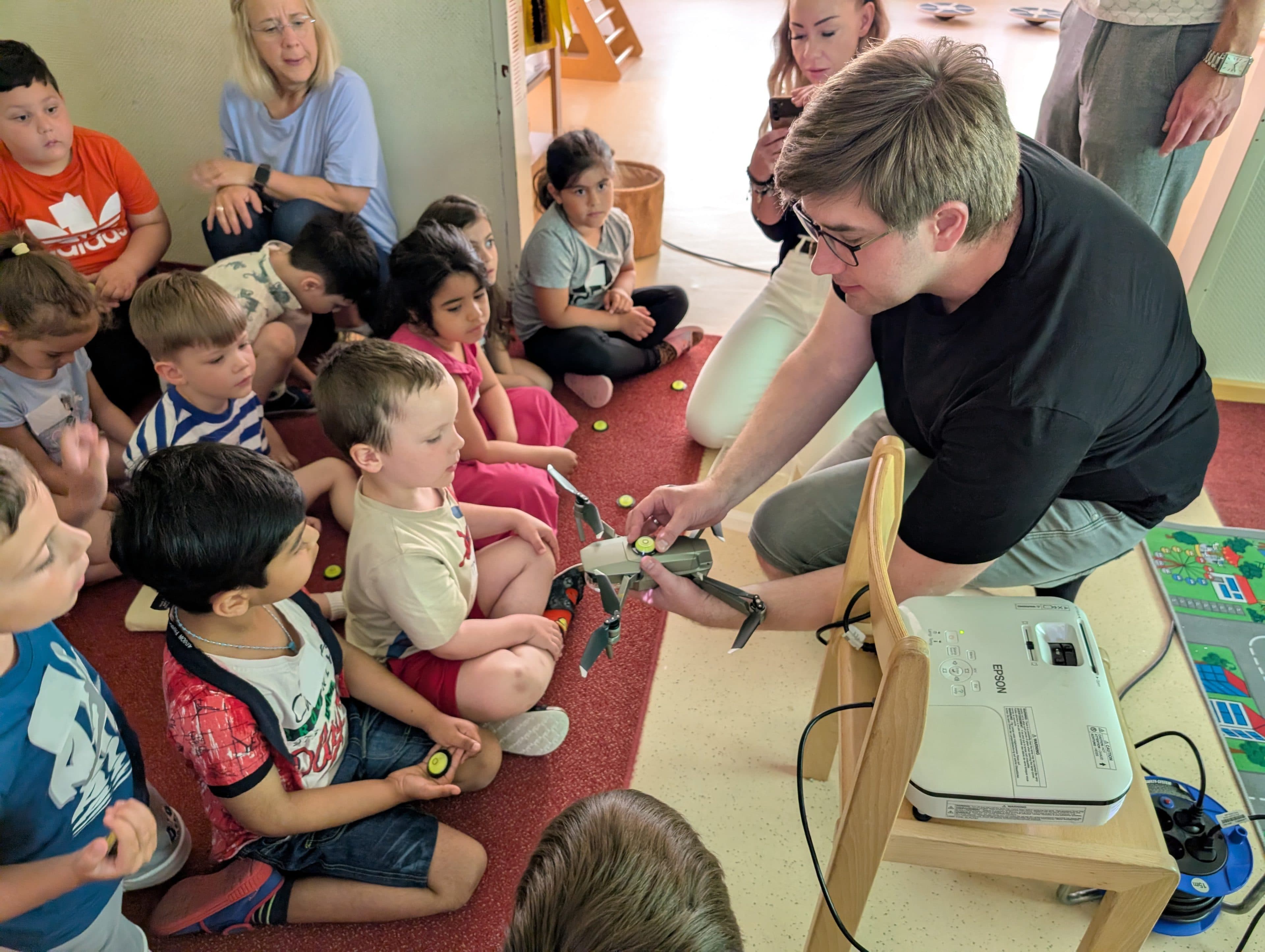 Eine Gruppe kleiner Kinder sitzt auf dem Boden um einen Erwachsenen herum, der eine kleine Drohne vorführt. Die Kinder schauen aufmerksam zu, einige halten gelbe Spielsteine in der Hand. In der Nähe sind ein Projektor und eine bunte Matte zu sehen.