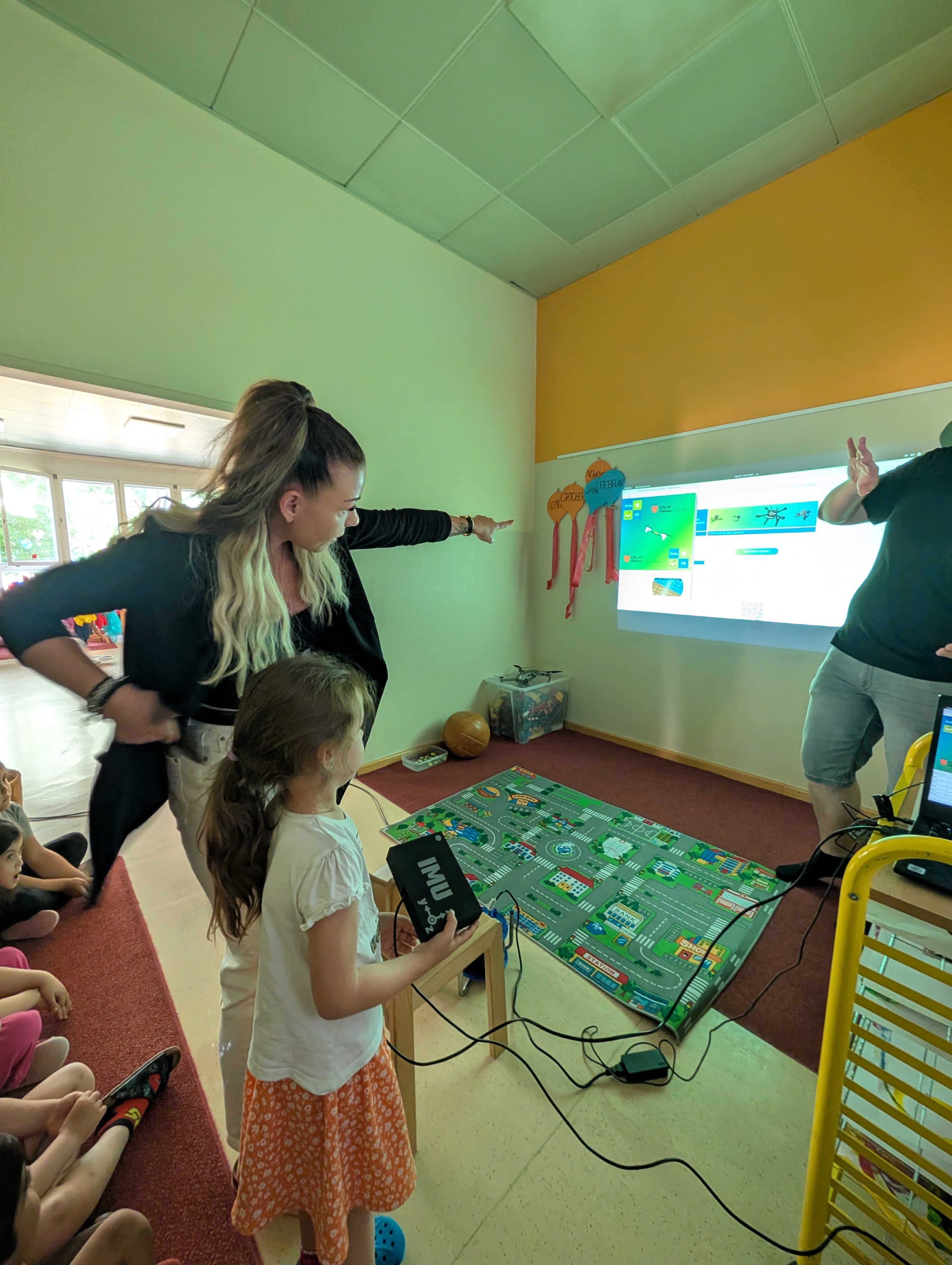 Eine Frau zeigt auf eine Leinwand an der Wand, während ein kleines Mädchen mit einer Fernbedienung in der Hand daneben steht. Kinder sitzen auf dem Boden und schauen zu. Der Raum ist bunt dekoriert, und auf dem Boden liegt eine Spielmatte.