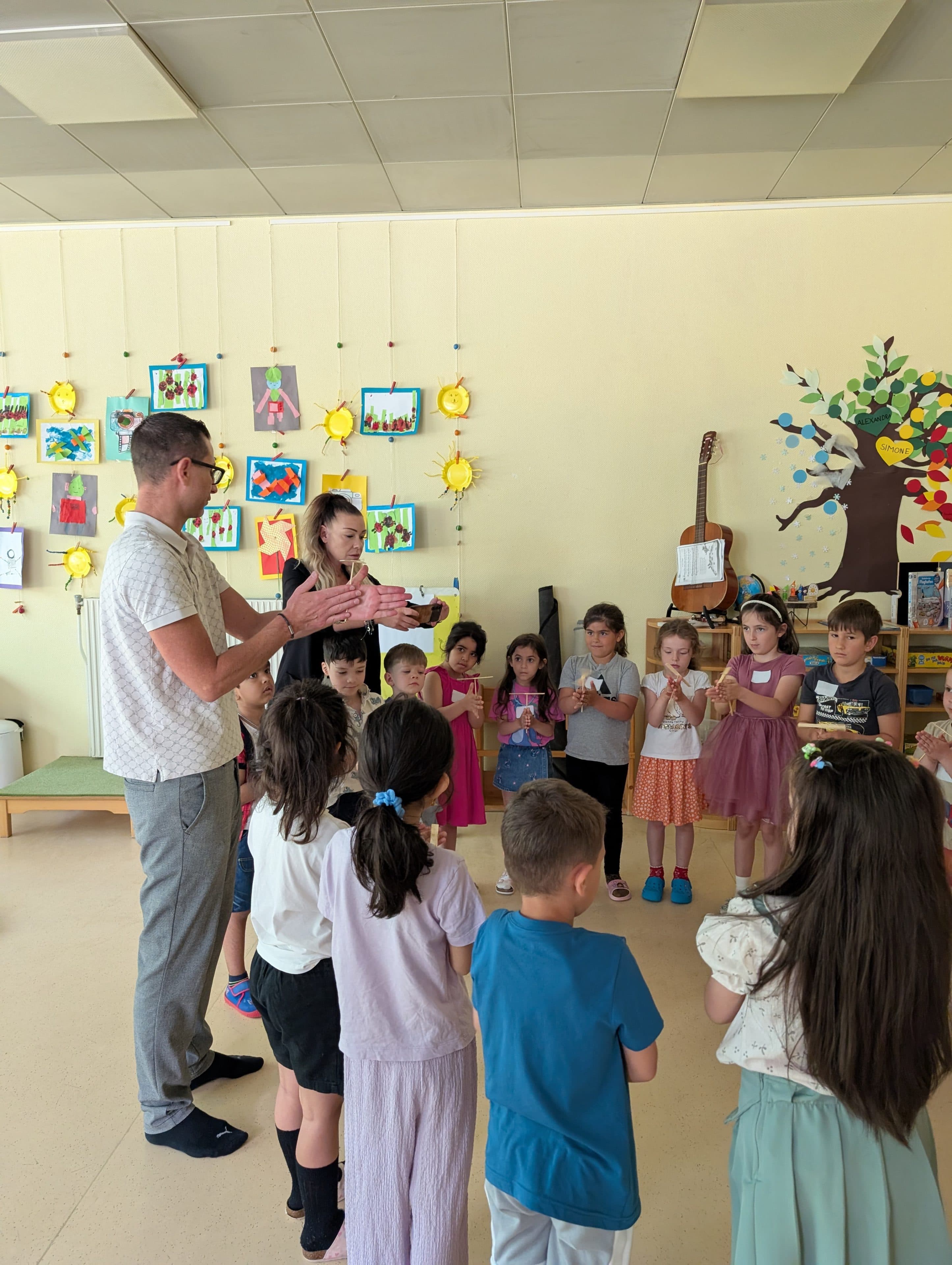 Eine Gruppe kleiner Kinder steht in einem Klassenzimmer im Kreis, während zwei Erwachsene eine Aktivität leiten. An den Wänden hängen bunte Bilder, Gitarren und Dekorationen. Die Kinder wirken interessiert und aufmerksam.