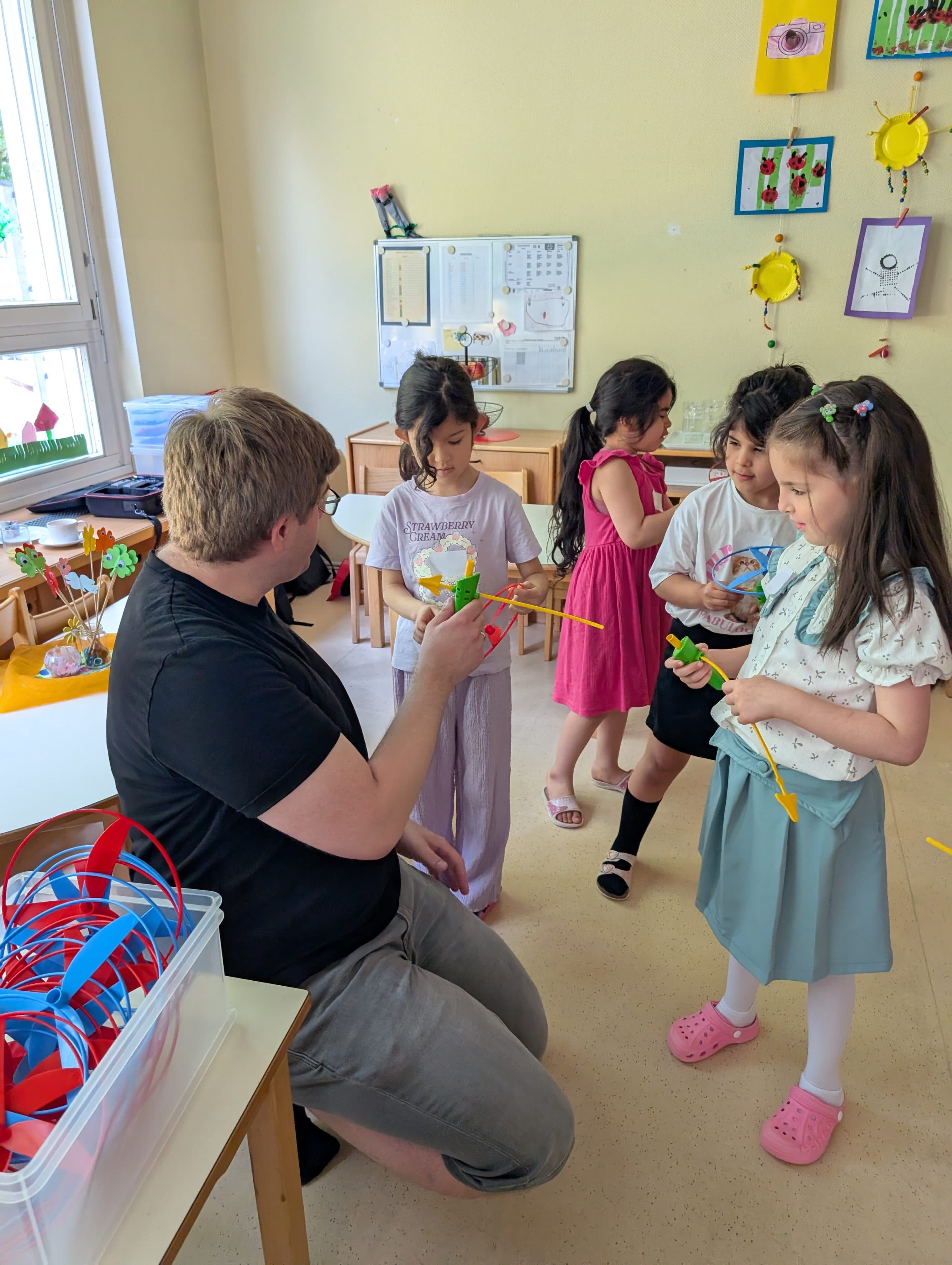 Ein Mann kniet in einem Klassenzimmer nieder und unterhält sich mit vier kleinen Mädchen, während er ihnen bunte Bastelarbeiten aushändigt. Die Kinder halten selbstgebastelte Blumendekorationen in den Händen und lächeln. An den Wänden sind Kunstwerke und Bastelarbeiten ausgestellt.