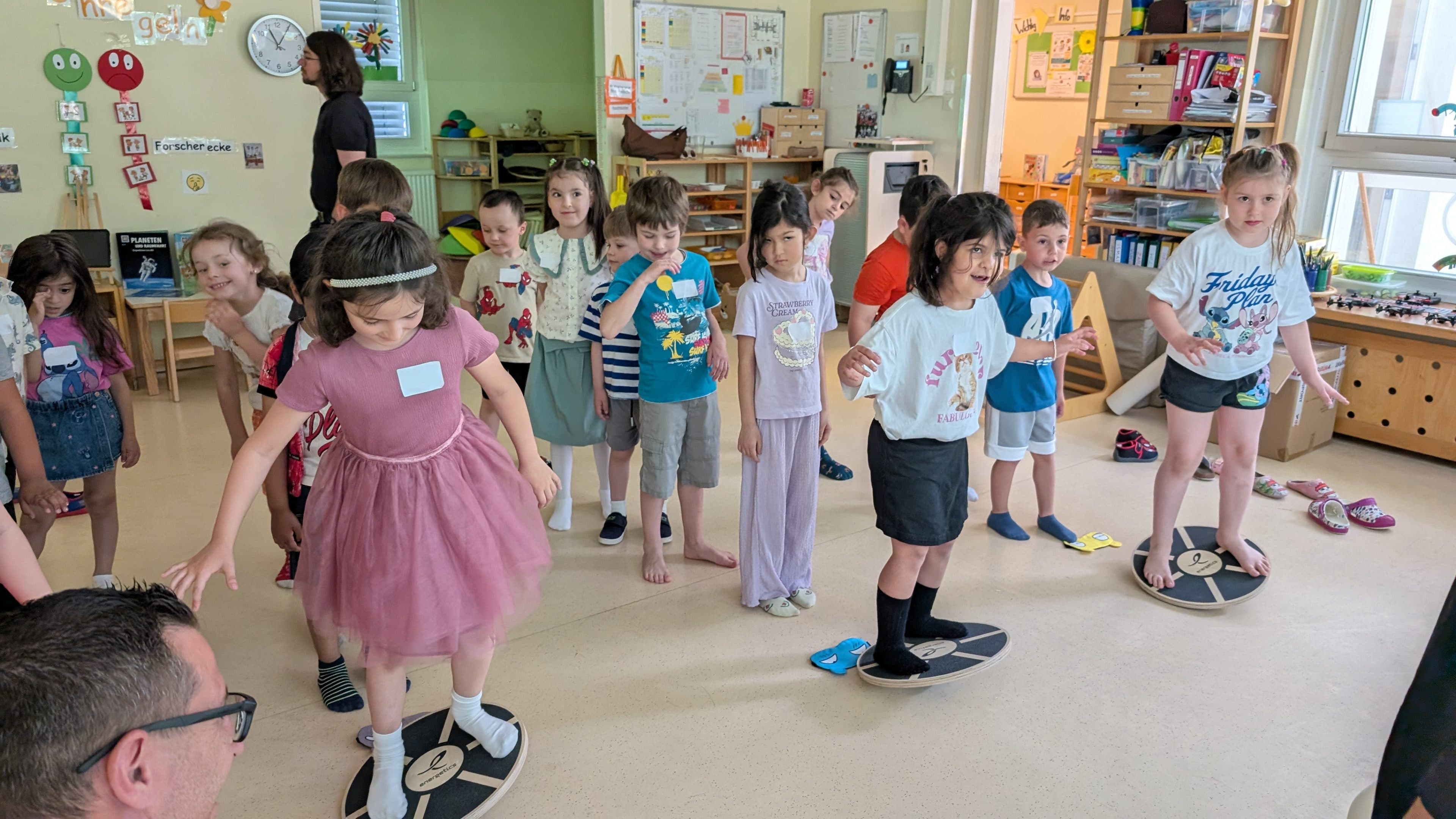 Eine Gruppe kleiner Kinder steht in einem Klassenzimmer auf Holzbrettern und balanciert darauf, dabei lächeln und lachen sie. Einige Kinder schauen zu, während andere sich abwechseln. Der Raum ist hell und farbenfroh, mit Lernplakaten und Regalen ausgestattet.