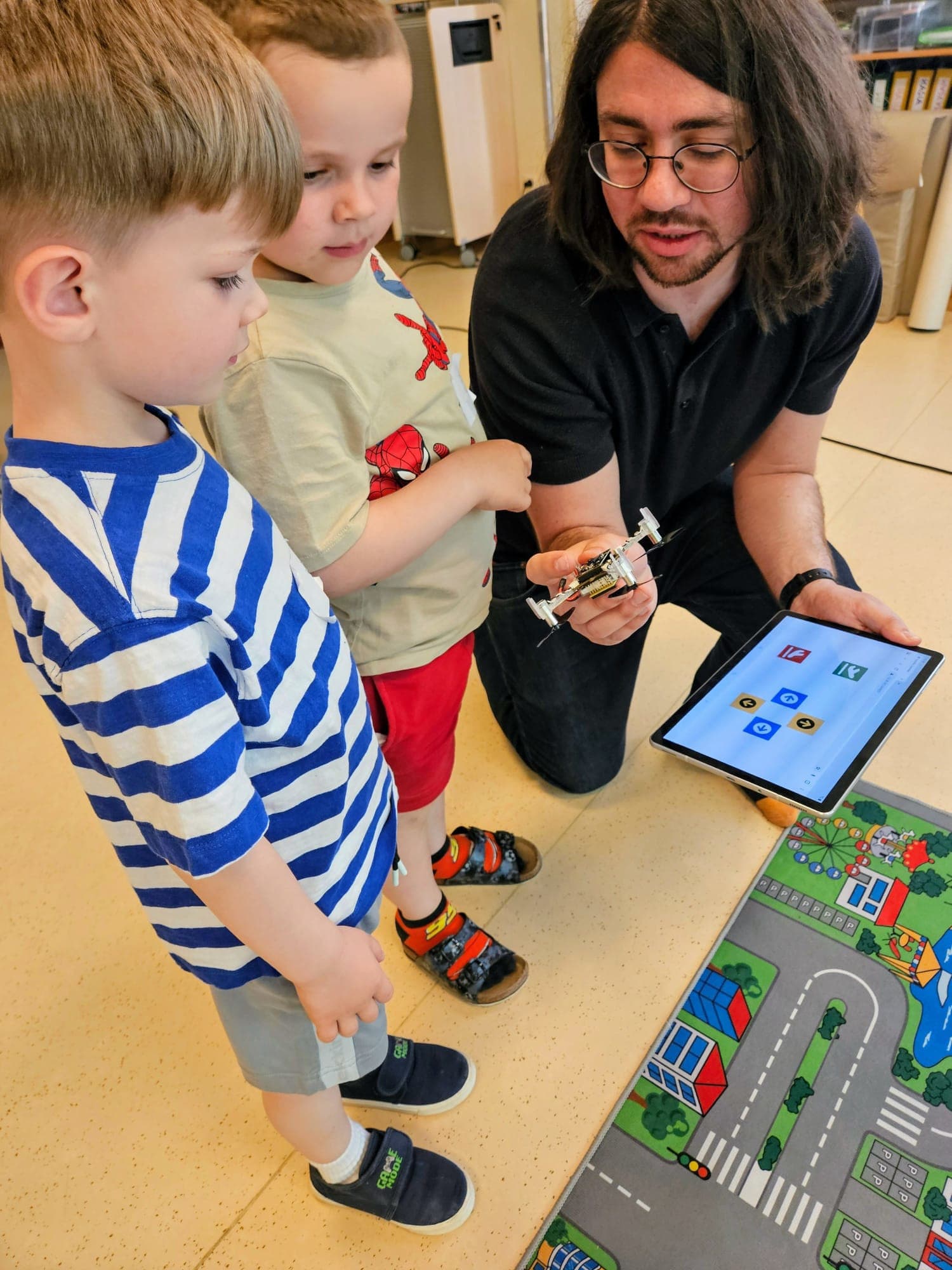 Ein Mann kniet neben zwei kleinen Jungen und zeigt ihnen eine kleine Drohne, während er ein Tablet mit bunten App-Symbolen in der Hand hält. Die Kinder schauen aufmerksam zu, während sie neben einer Spielmatte mit Straßen und Gebäuden sitzen.