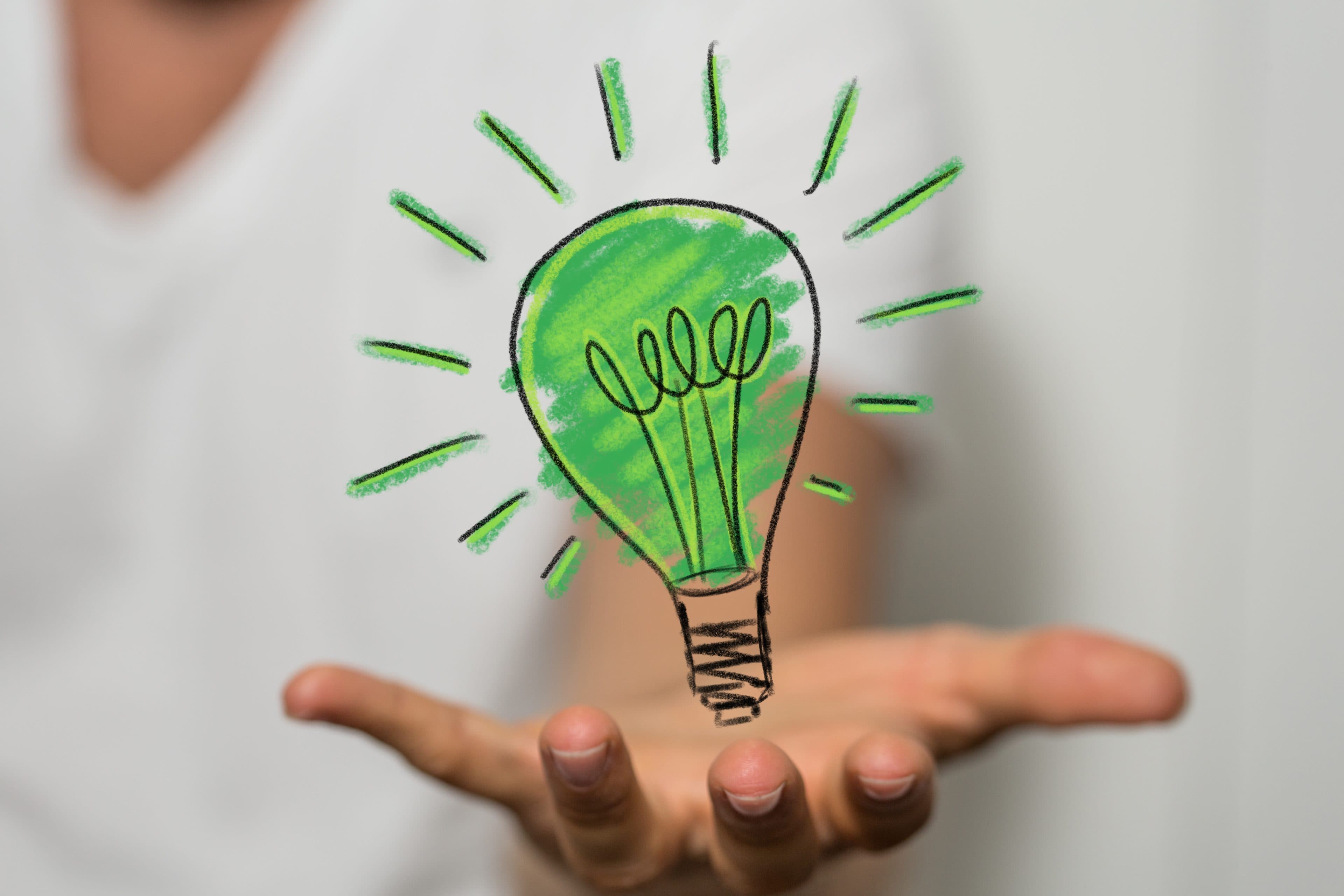 Eine Person streckt ihre Hand mit der Handfläche nach oben aus; darüber sind digital eine grüne Glühbirne und leuchtende Strahlen dargestellt, die für Kreativität oder umweltfreundliche Ideen stehen.