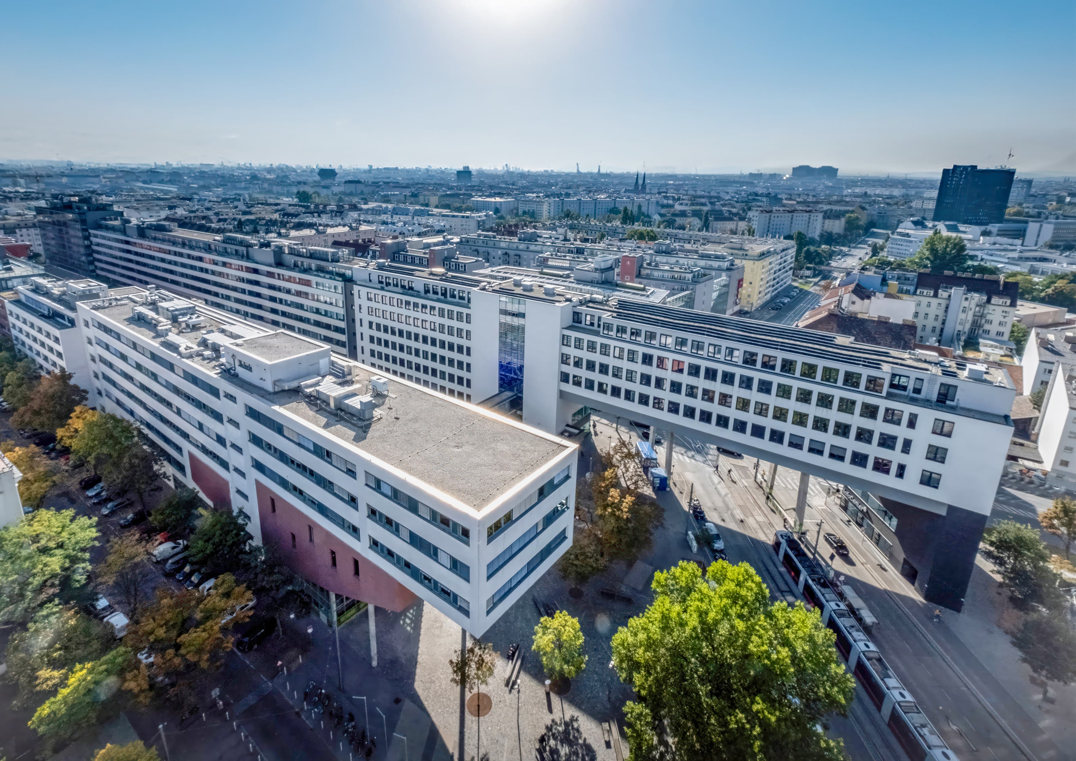 Luftaufnahme eines großen, modernen Gebäudes mit weißen Fassaden und geometrischen Formen, das eine Straßenbahnlinie überspannt – in einer städtischen Kulisse mit Bäumen und in der Ferne aufragenden Wolkenkratzern unter einem strahlend blauen Himmel.