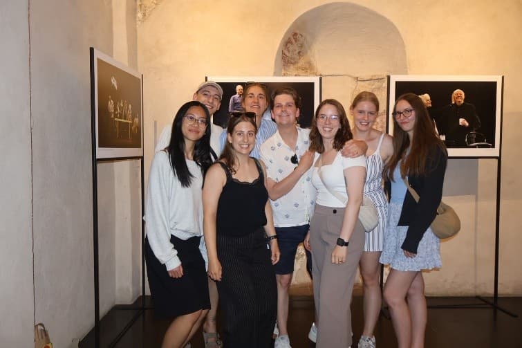 Eine Gruppe von acht jungen Erwachsenen posiert gemeinsam in einem Innenraum vor zwei gerahmten Fotos und lächelt in lässiger Sommerkleidung, dicht beieinander stehend. Die Kulisse besteht aus Steinwänden und einer gewölbten Nische.