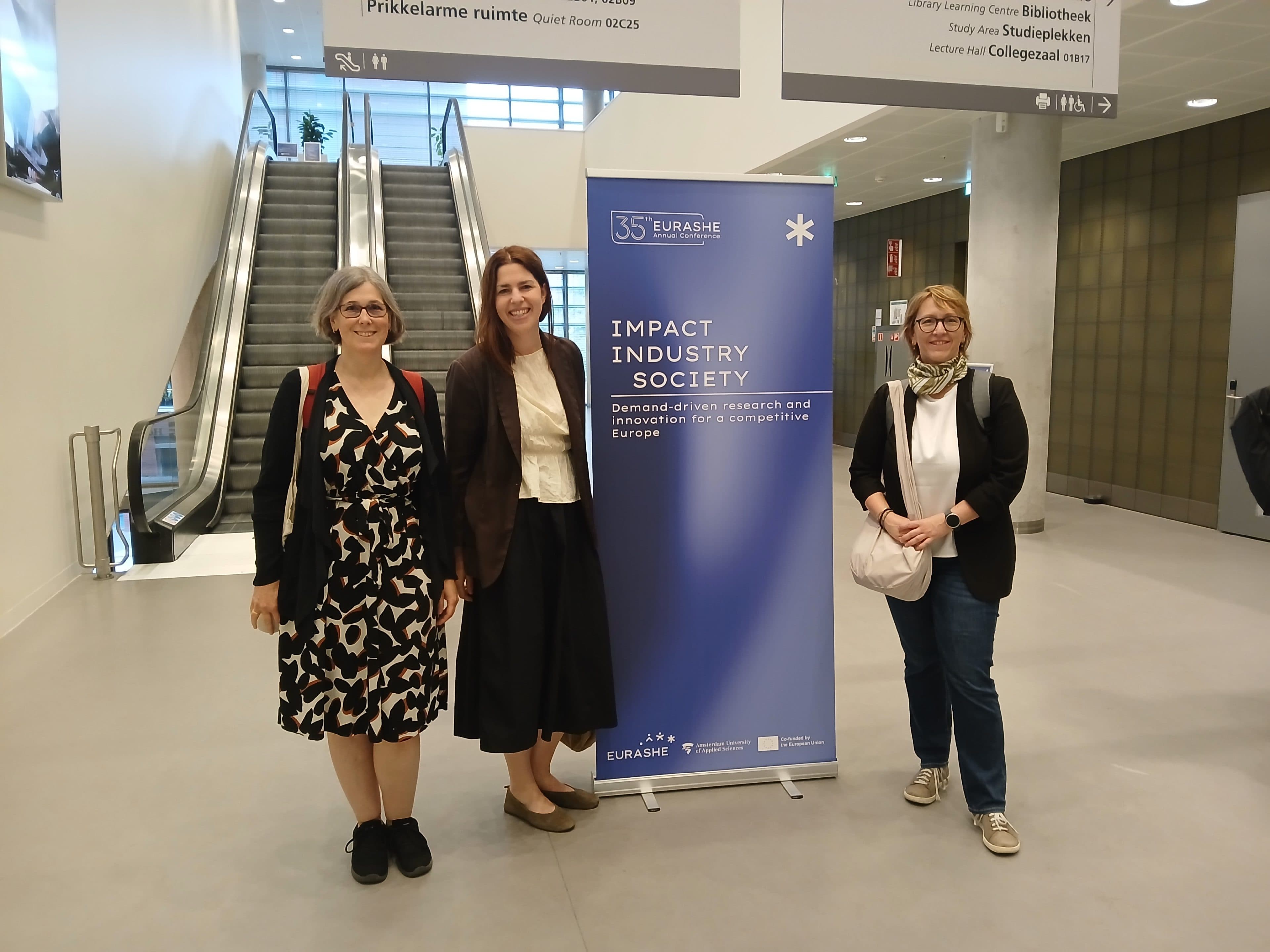 Drei Frauen stehen lächelnd neben einem blauen Banner mit der Aufschrift „Impact, Industry, Society“ in einem Konferenzsaal, im Hintergrund sind eine Rolltreppe und Wegweiser zu sehen.