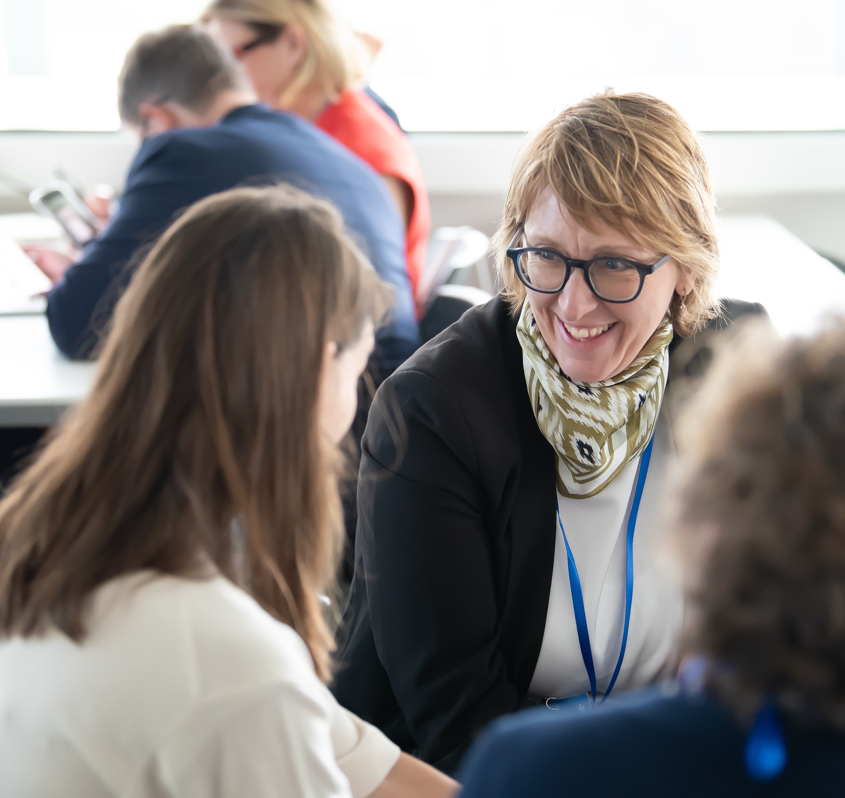 Zwei Frauen lächeln und unterhalten sich in einem hellen Raum im Rahmen einer Veranstaltung, die offenbar beruflicher Natur ist oder dem Networking dient, während sich im Hintergrund andere Personen unterhalten.