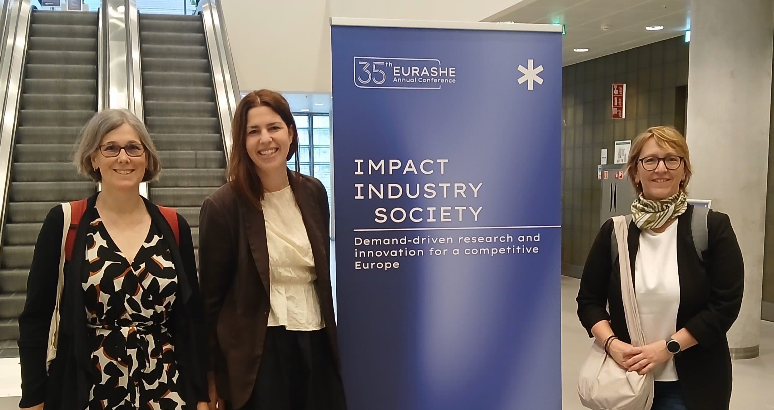 Drei Frauen stehen lächelnd neben einem blauen Plakat mit der Aufschrift „Impact Industry Society“ in einem Veranstaltungsraum in der Nähe einer Rolltreppe. Die Umgebung ist modern gestaltet, mit Schildern und einem Fliesenboden.