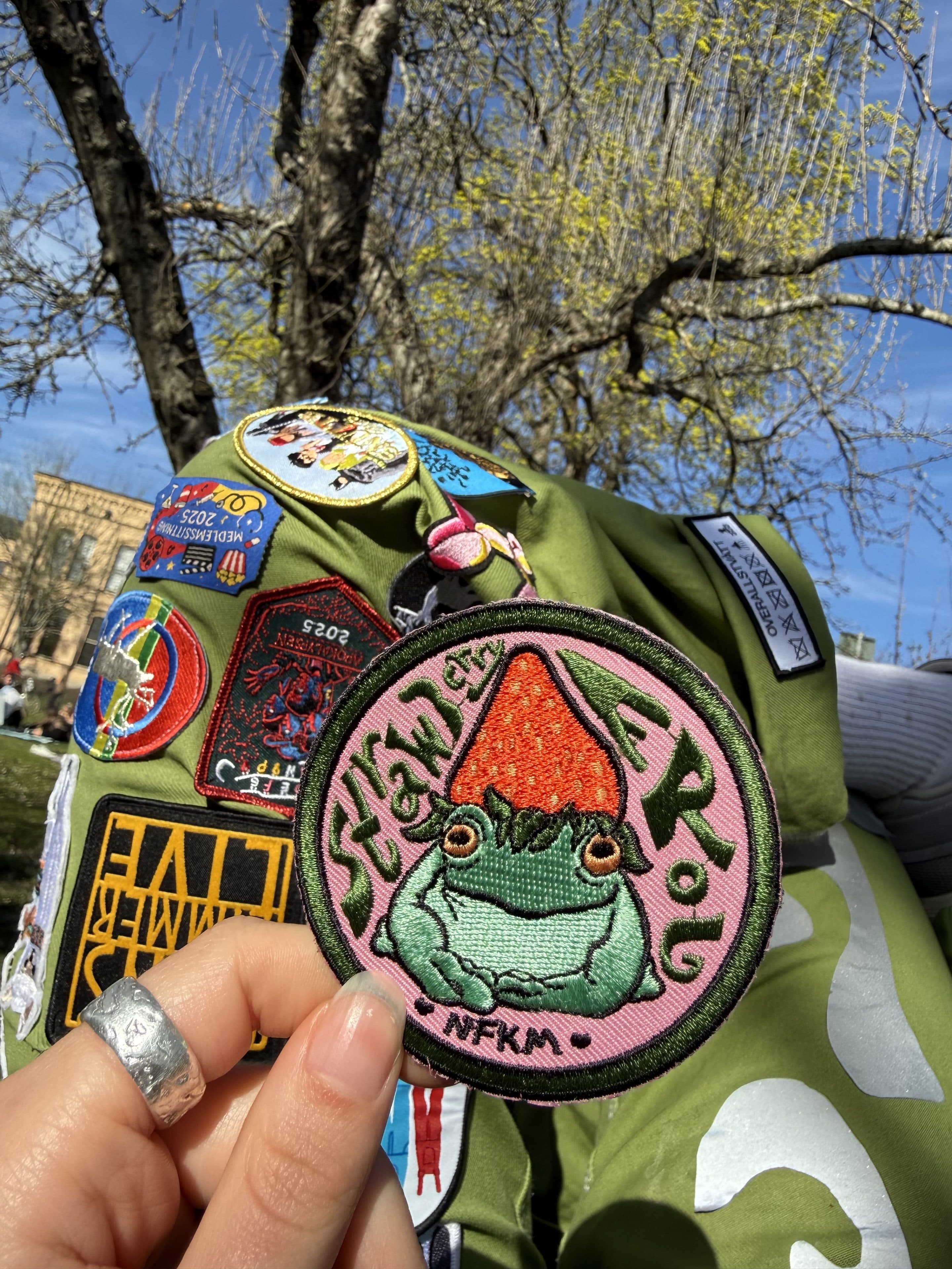 Eine Hand hält einen runden, bestickten Aufnäher, auf dem ein Frosch mit einem roten Pilzhut und die Aufschrift „Setäsi on ERoL, NFKM“ zu sehen ist. Der Aufnäher ist an einer grünen Jacke mit verschiedenen bunten Aufnähern befestigt. Im Hintergrund sind Bäume und ein blauer Himmel zu sehen.