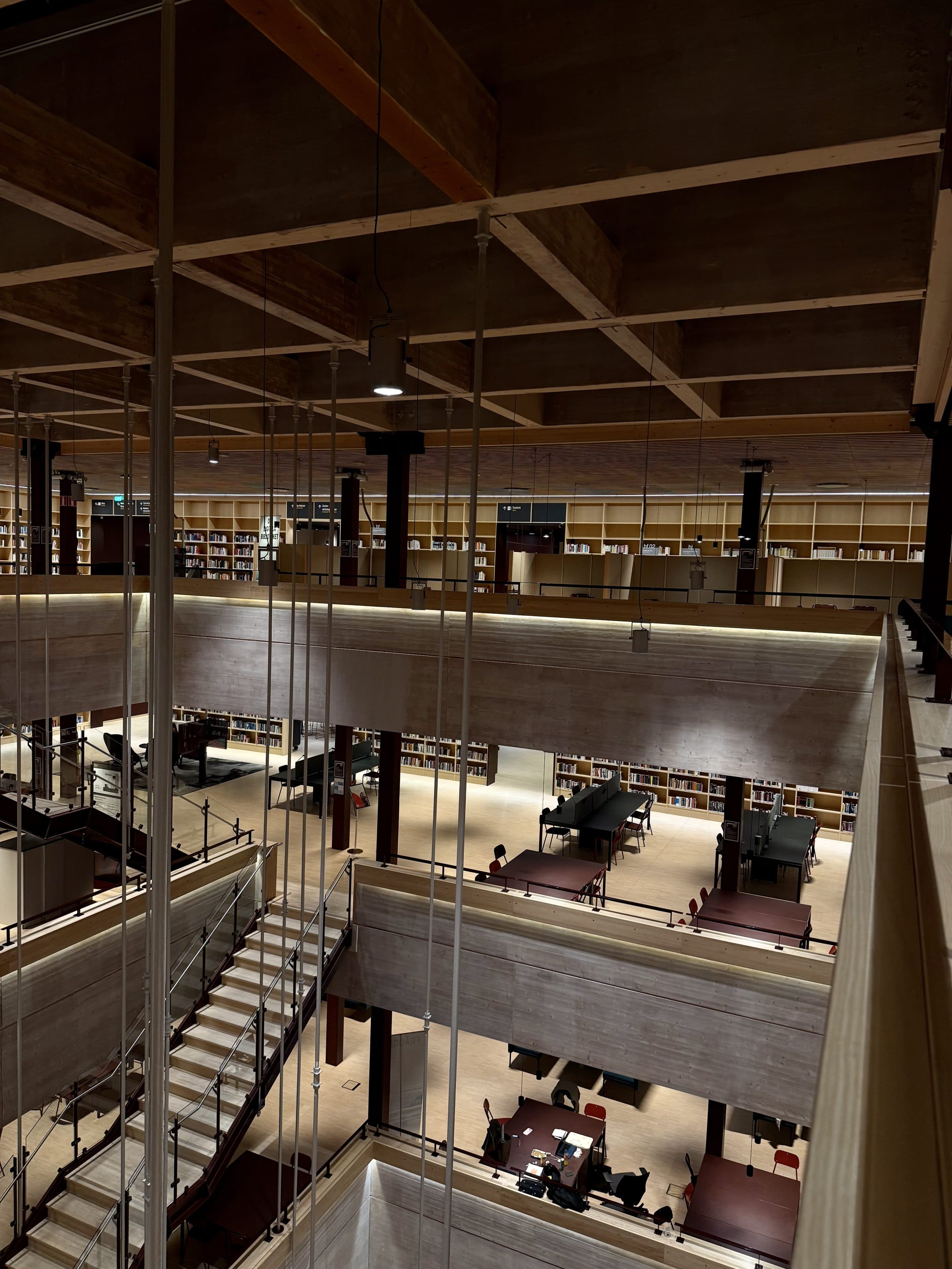 Eine geräumige, moderne Bibliothek mit mehreren Etagen, Bücherregalen an den Wänden und Lerntischen, an denen Menschen sitzen. Sichtbare Betonbalken und Holzakzente sind zu sehen, ebenso wie eine zentrale Treppe, die die Etagen miteinander verbindet.