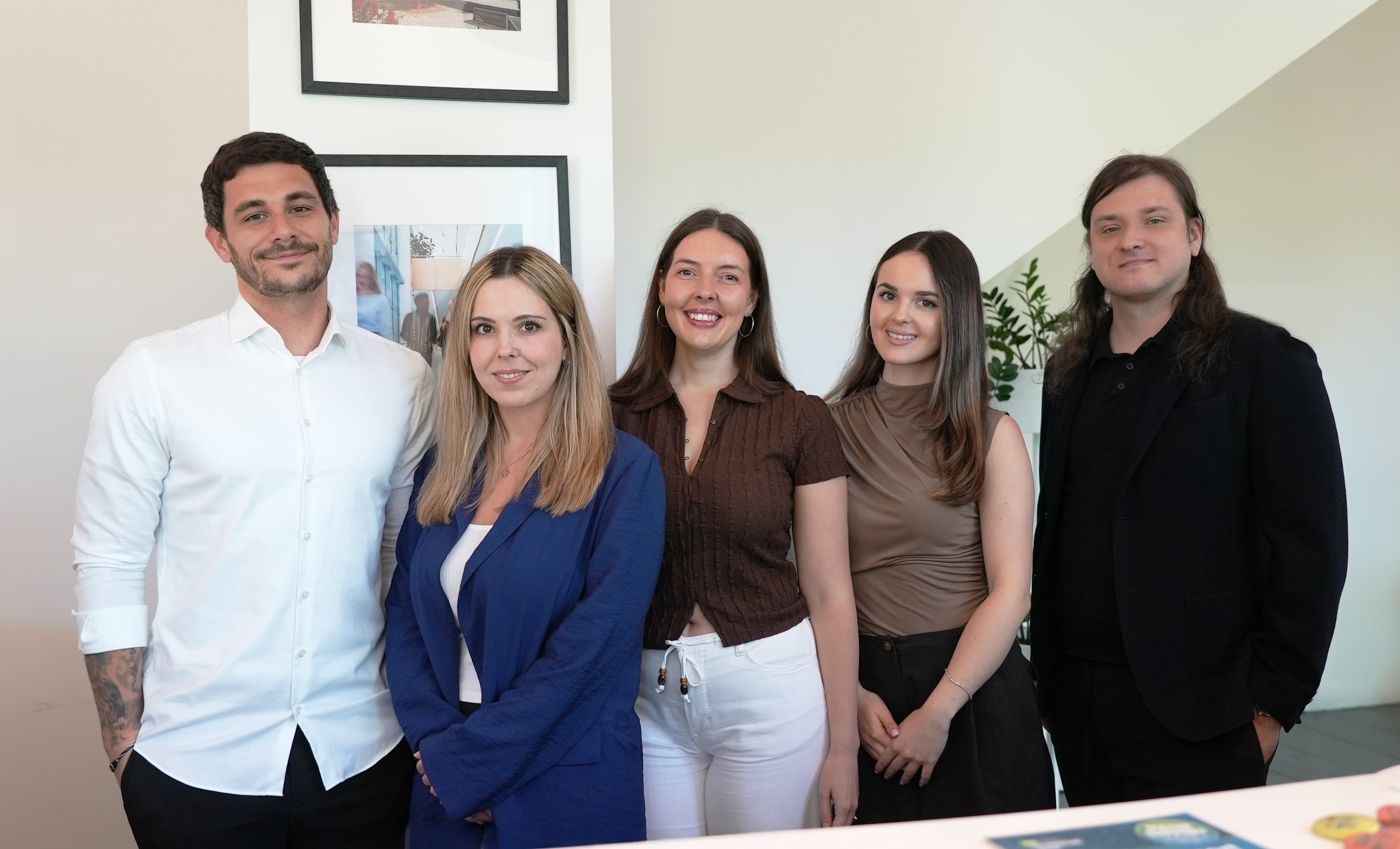 Fünf Personen stehen in einem Raum und lächeln in die Kamera. In der Mitte stehen drei Frauen, flankiert von zwei Männern. Sie tragen Business-Casual-Kleidung; im Hintergrund sind gerahmte Fotos und eine Pflanze zu sehen.