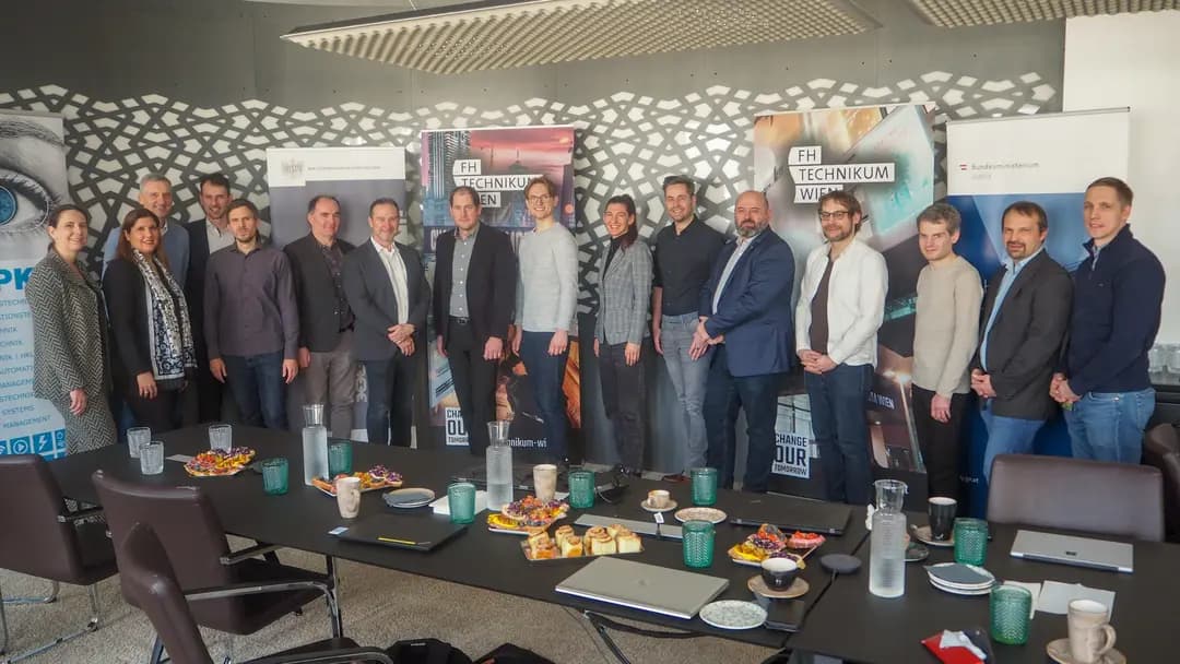 Eine Gruppe von sechzehn Personen posiert für ein Foto in einem Konferenzraum, in dem Banner, Snacks, Getränke und Unterlagen auf dem Tisch liegen. Im Hintergrund sind eine moderne Einrichtung und Banner des FH Technikum Wien zu sehen.
