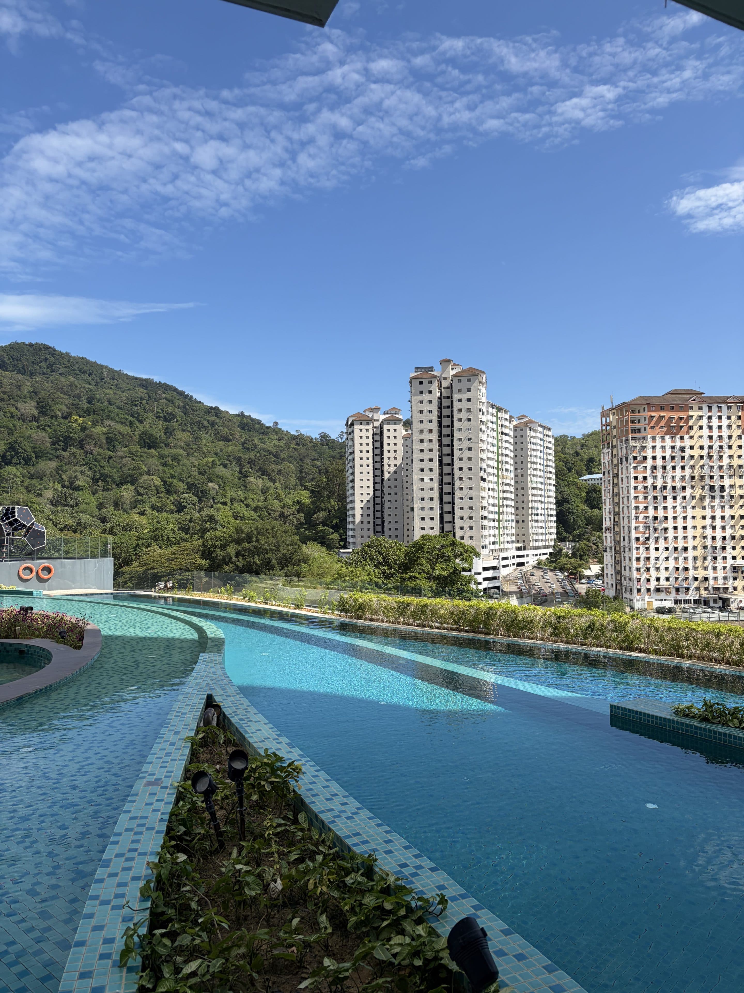 Ein Infinity-Pool auf dem Dach mit kristallklarem, blauem Wasser bietet unter einem strahlenden, teilweise bewölkten Himmel einen Blick auf grüne Hügel und hohe Wohnhochhäuser.