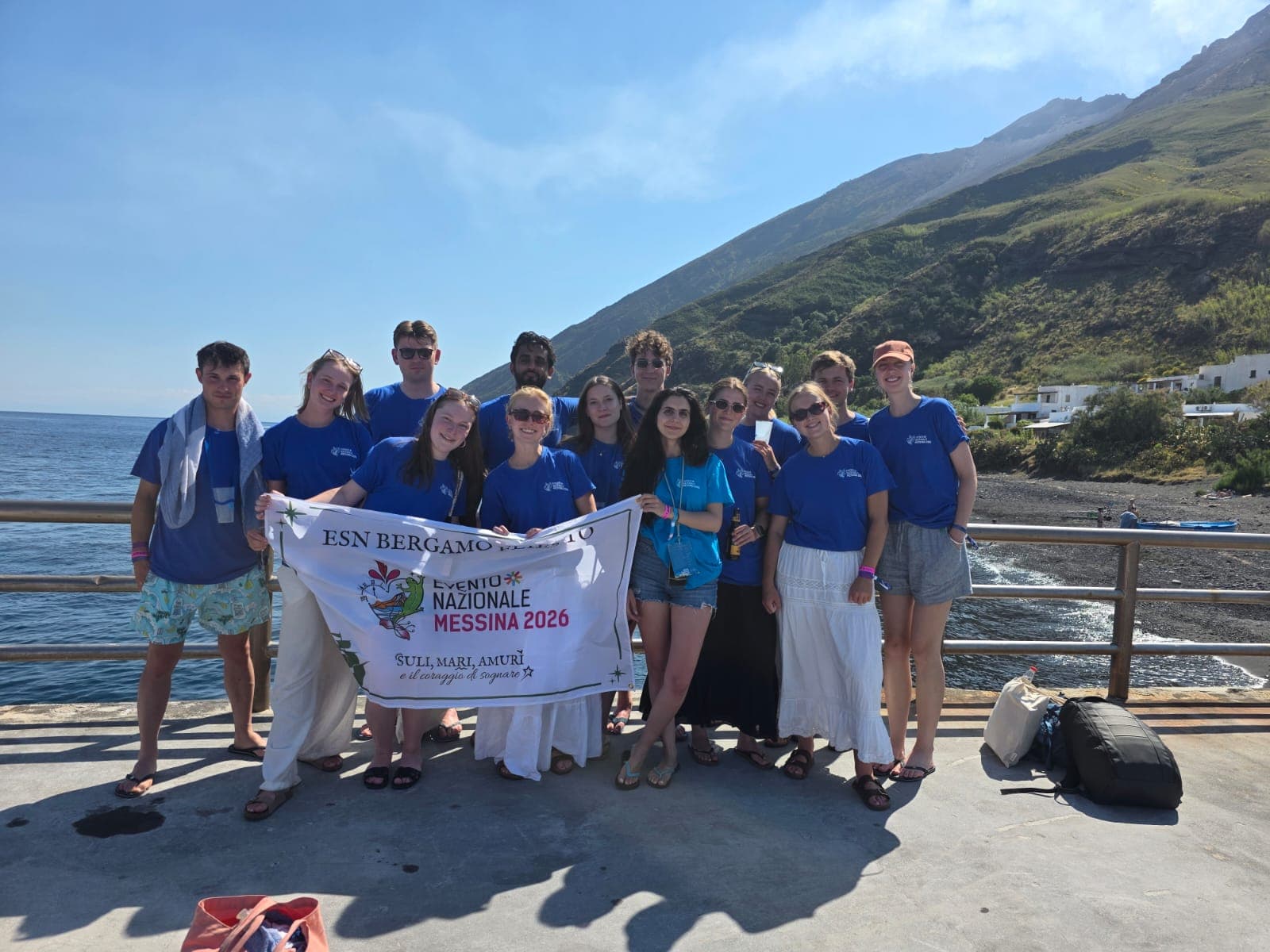 Eine Gruppe von Menschen in blauen T-Shirts steht auf einem Steg am Meer, lächelt und hält ein Transparent mit der Aufschrift „ESN Bergamo Evento Nazionale Messina 2026“ in die Höhe, im Hintergrund sind Berge und Häuser zu sehen.