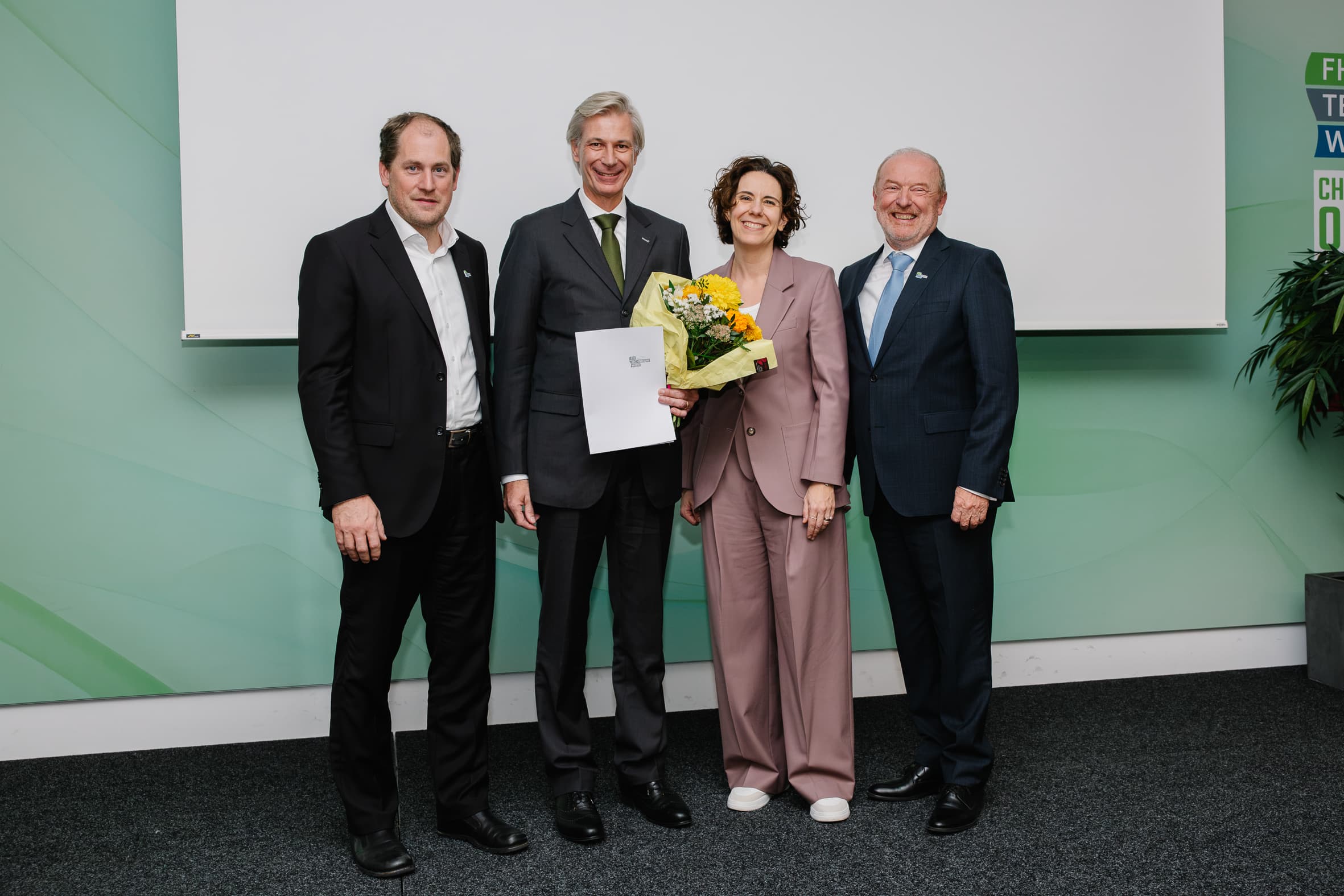 Vier Personen stehen bei einer beruflichen Veranstaltung nebeneinander in einer Reihe, wobei die Frau in der Mitte einen Strauß gelber Blumen und ein Dokument in der Hand hält.