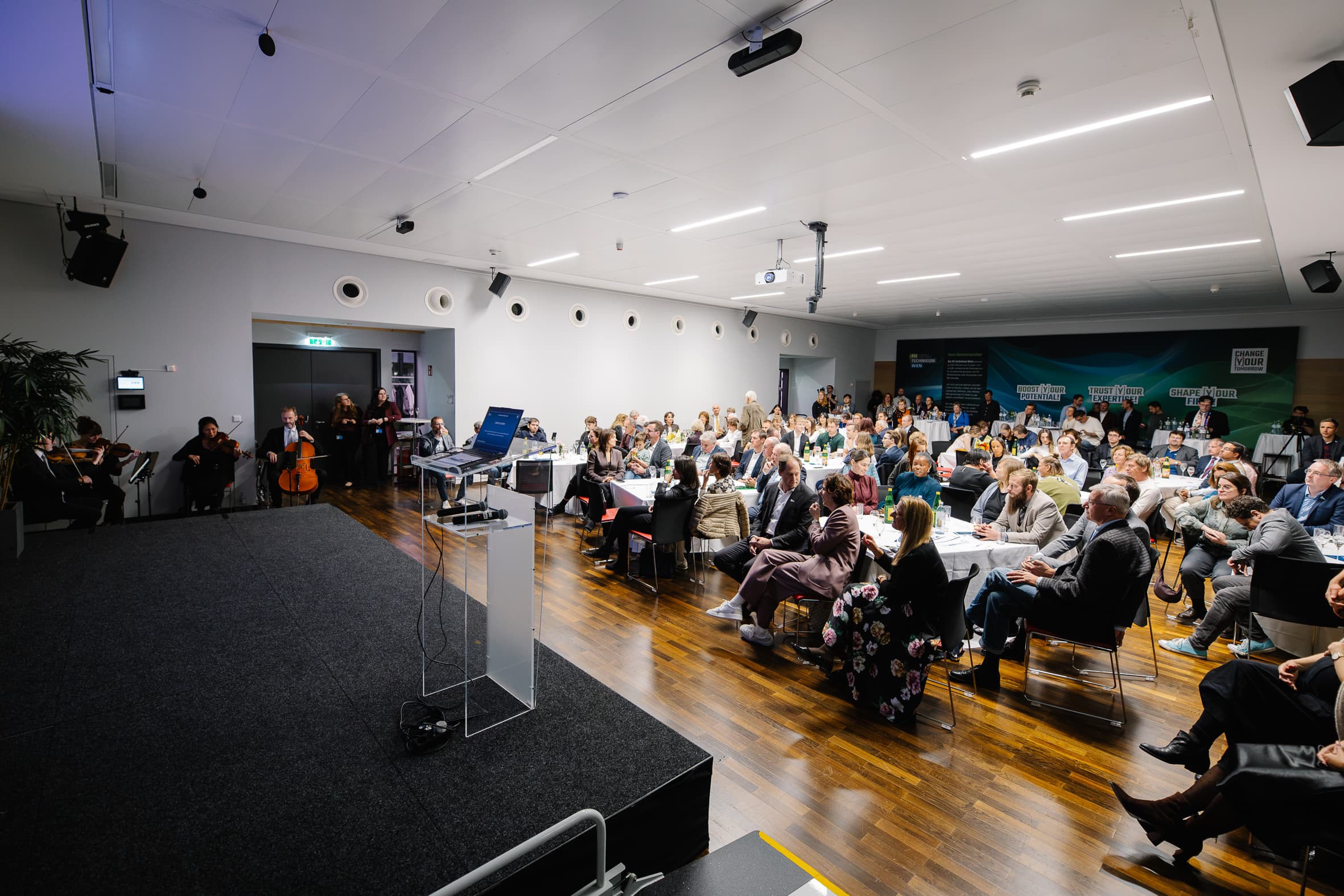 Ein kleines Ensemble spielt auf einer Bühne Streichinstrumente vor einem Publikum, das an Tischen in einem großen Konferenzraum sitzt.