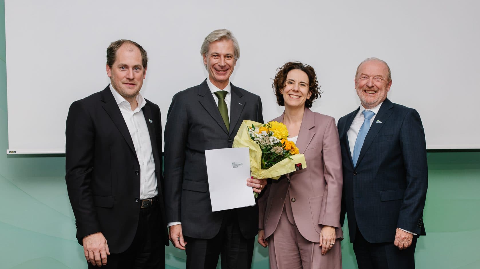Vier Personen stehen für ein Foto in einer Reihe, wobei die zweite Person von links ein Dokument und einen Blumenstrauß in der Hand hält.