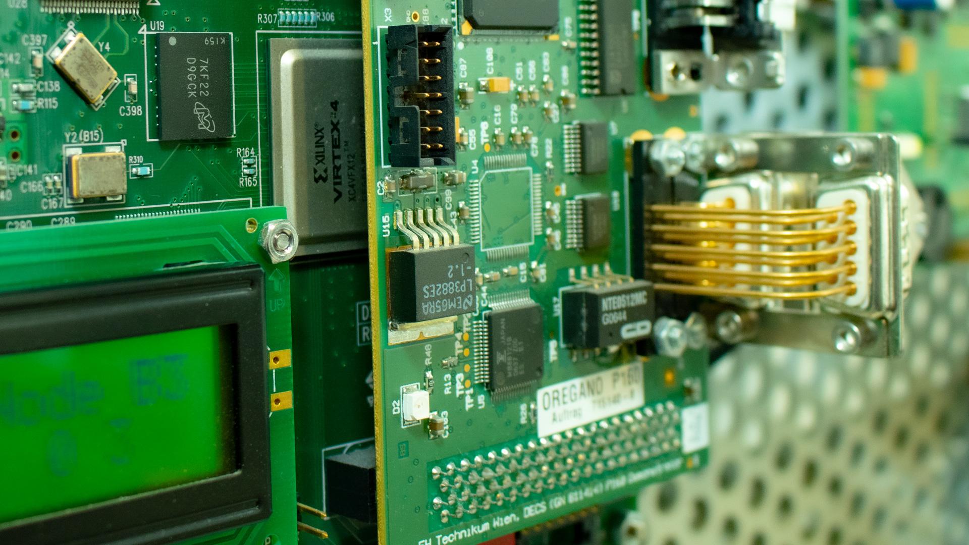 Nahaufnahme einer grünen Leiterplatte mit verschiedenen Mikrochips, elektronischen Bauteilen, einer digitalen Anzeige und goldenen Drähten, die mit einem Stecker auf der rechten Seite verbunden sind. Im Hintergrund ist ein Teil einer perforierten Metalloberfläche zu sehen.