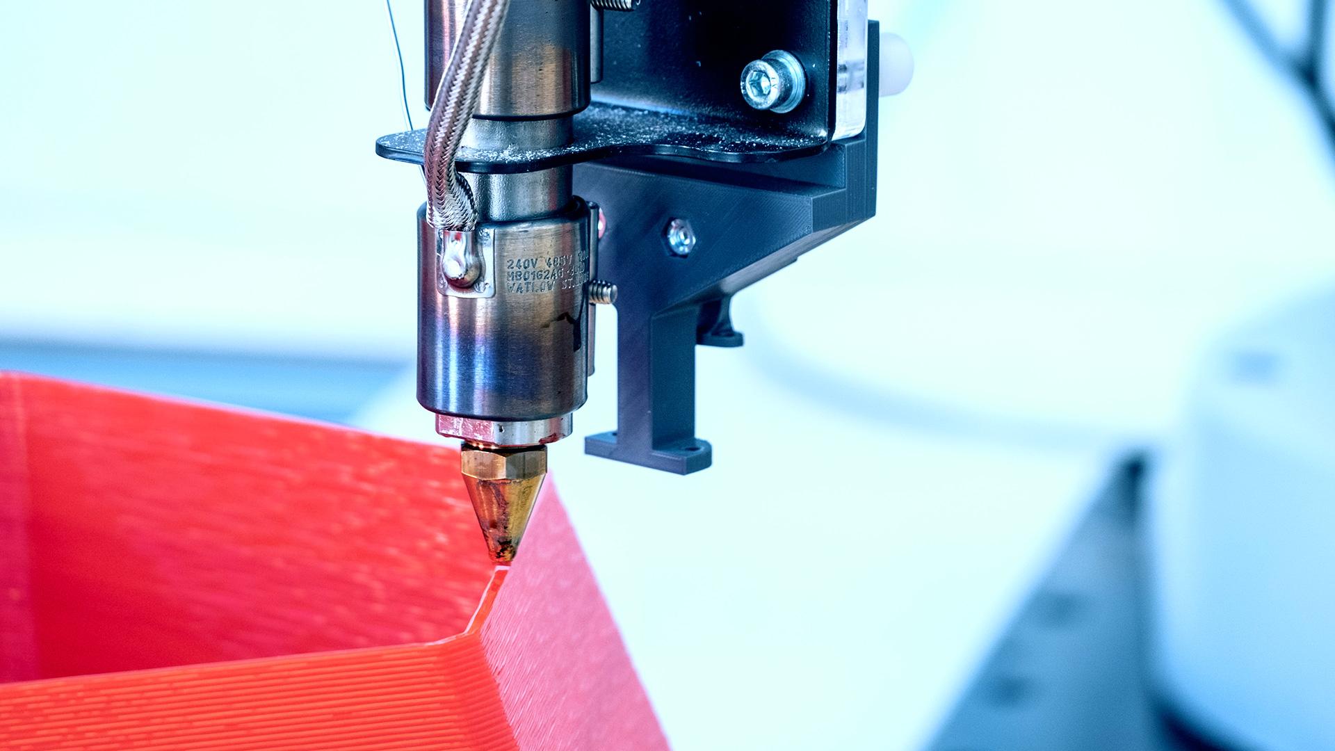 Nahaufnahme einer 3D-Druckerdüse, die rotes Filamentmaterial aufträgt, um ein geometrisches Objekt zu erstellen, wobei im Hintergrund mechanische Teile und Kabel zu sehen sind.
