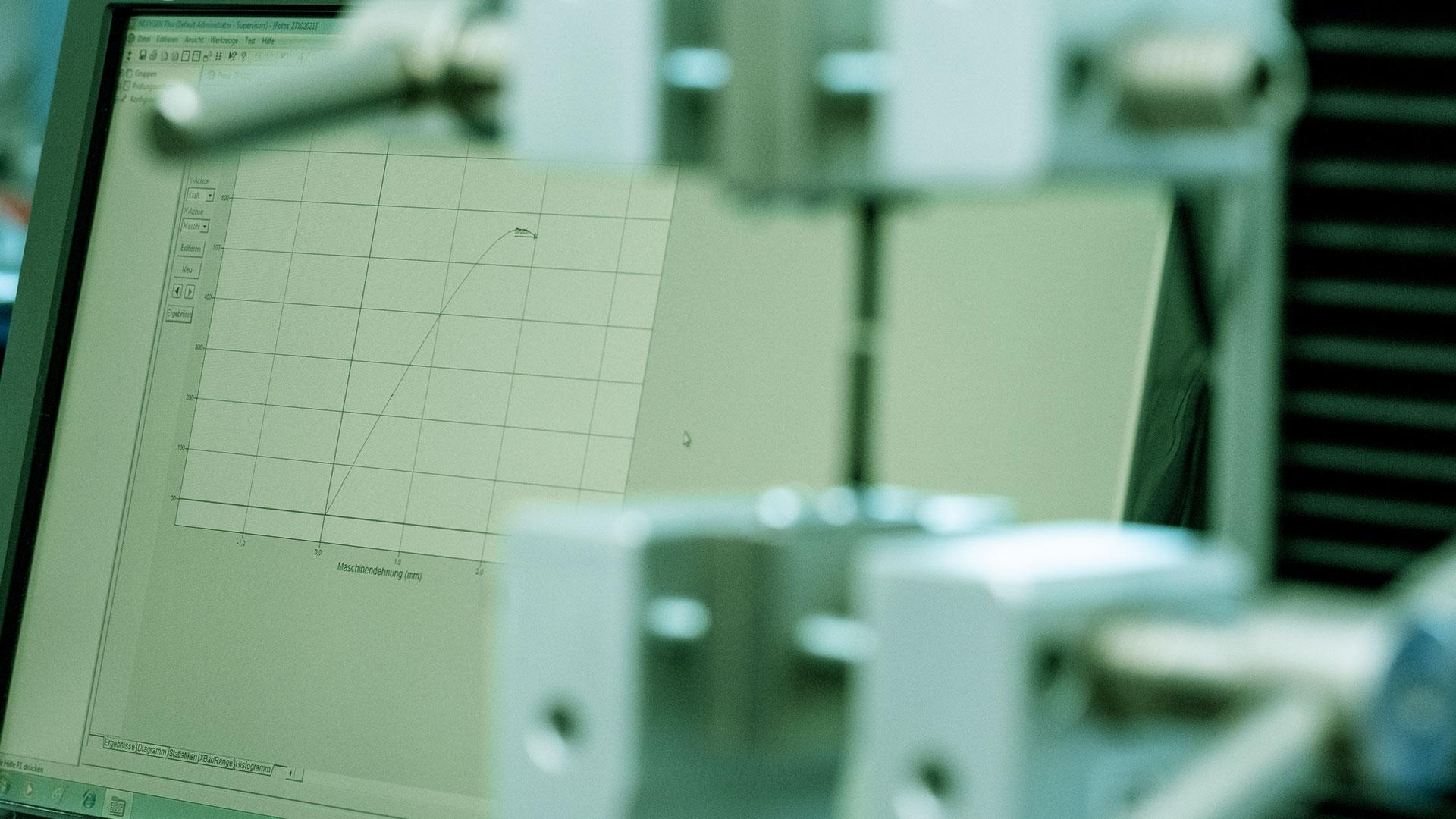 Nahaufnahme einer Zugprüfmaschine, die eine Probe einspannt, mit einem Computerbildschirm im Hintergrund, auf dem ein Diagramm mit den Spannungs-/Dehnungsdaten angezeigt wird.