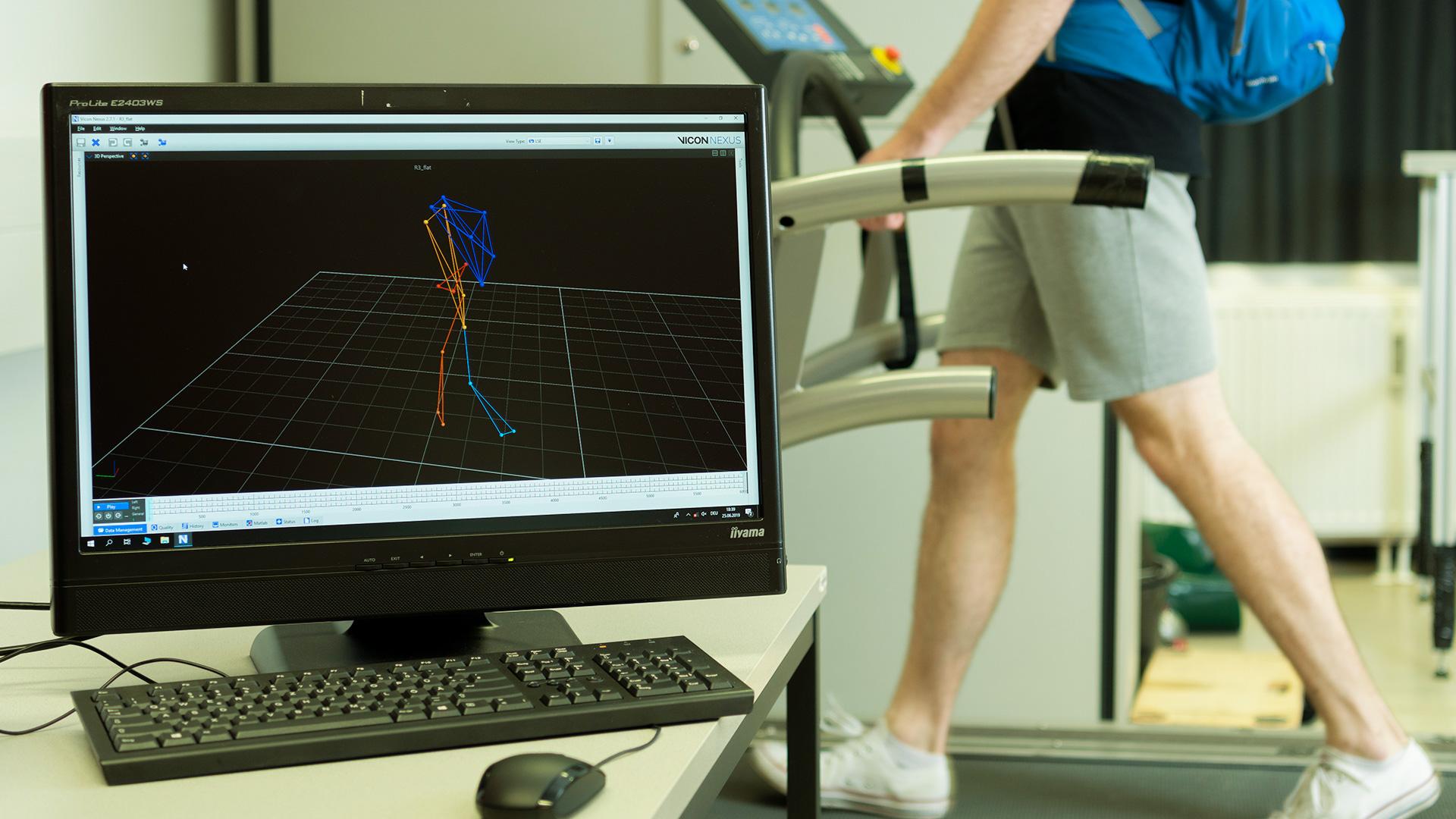 Auf einem Computermonitor wird ein 3D-Bewegungserfassungsmodell angezeigt, während im Hintergrund eine Person in Shorts und mit blauem Rucksack auf einem Laufband läuft.
