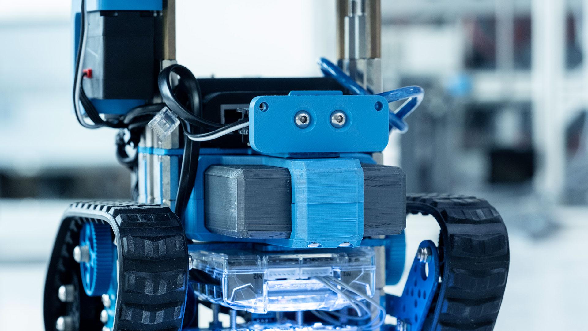 Nahaufnahme eines blauen und grauen Raupenroboters auf weißem Hintergrund, der seine Räder, Sensoren und freiliegenden internen Komponenten mit Drähten und durchsichtigem Gehäuse zeigt.