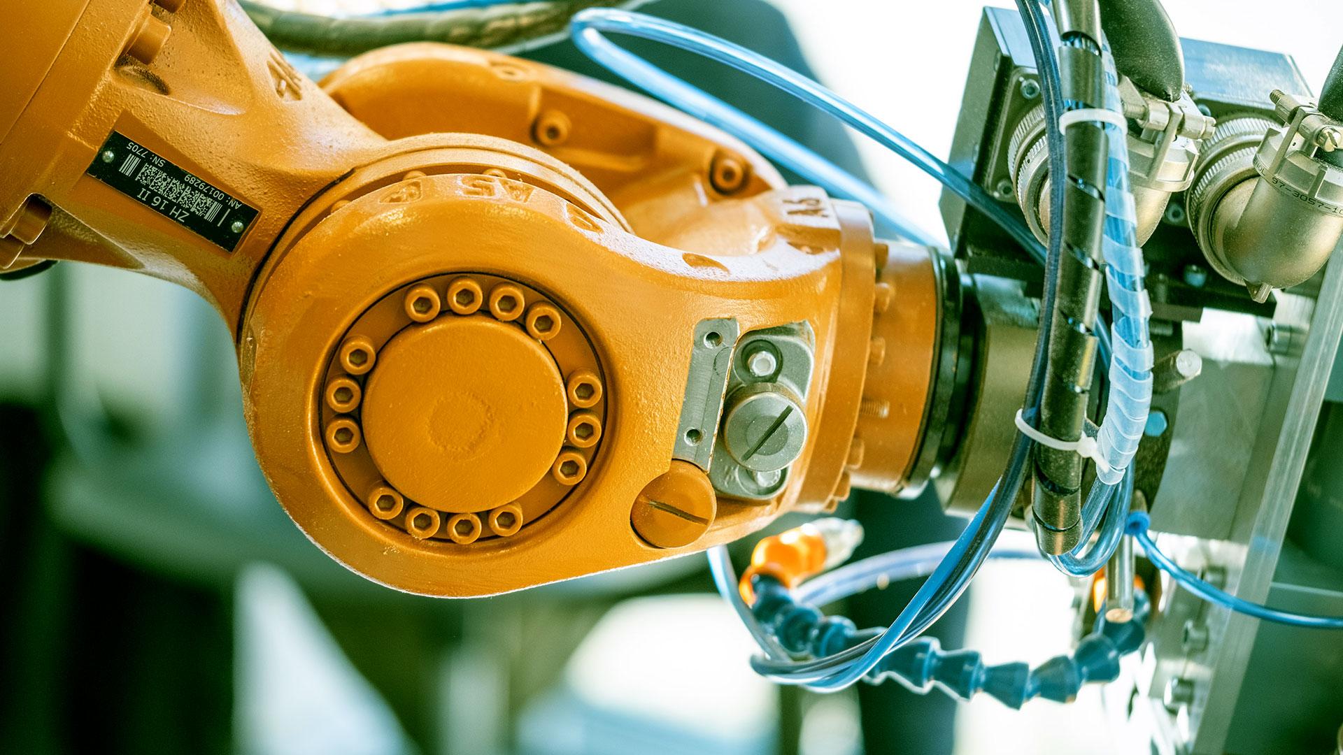 Nahaufnahme eines orangefarbenen Industrieroboterarms mit sichtbaren Schrauben und Kabeln, der mit Maschinen verbunden ist und mechanische Teile und Automatisierungstechnik hervorhebt.