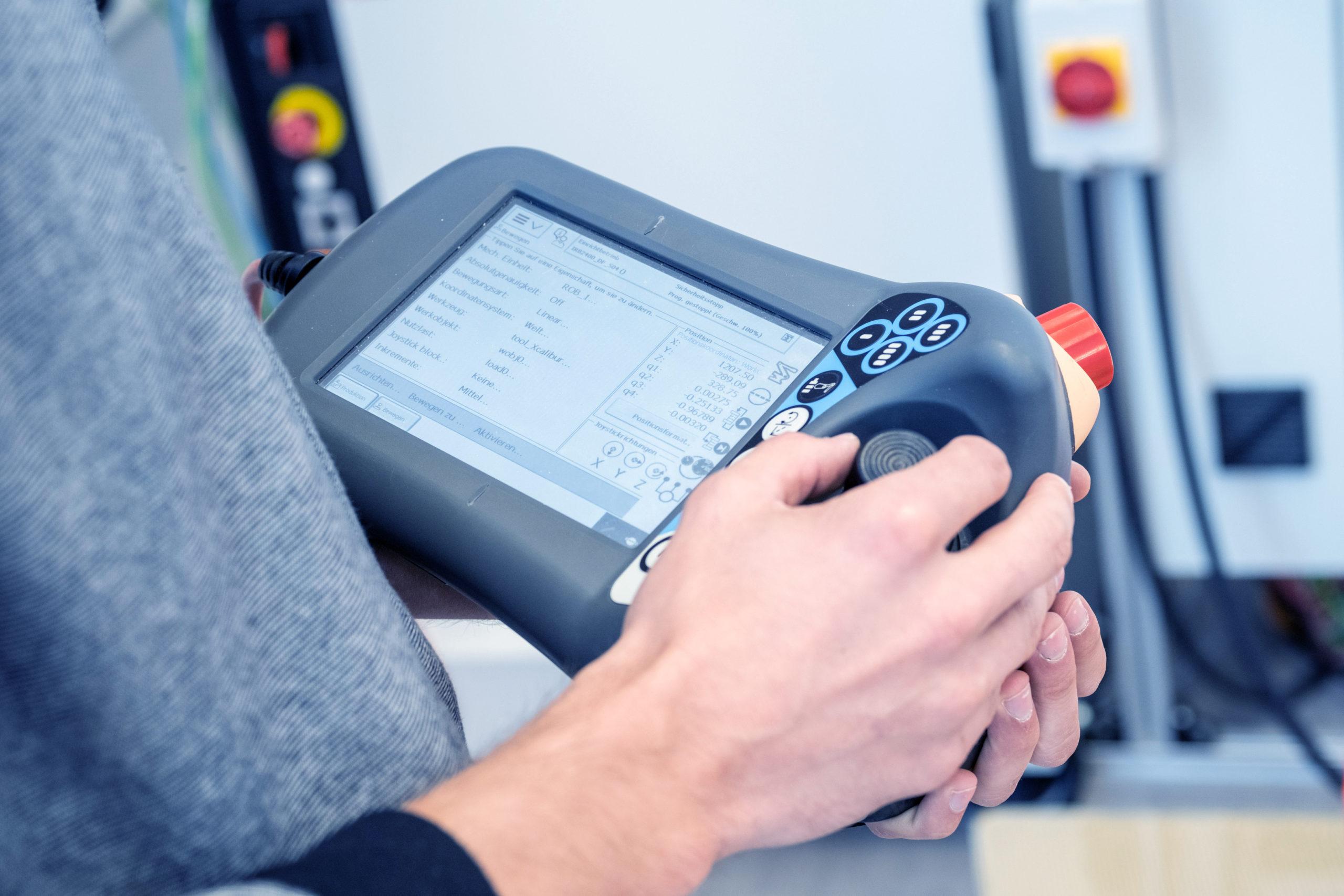 Eine Person, die ein handgehaltenes industrielles Bedienfeld oder eine Fernbedienung mit einem Bildschirm und Tasten hält und bedient, das wahrscheinlich zur Programmierung oder Steuerung von Maschinen verwendet wird.