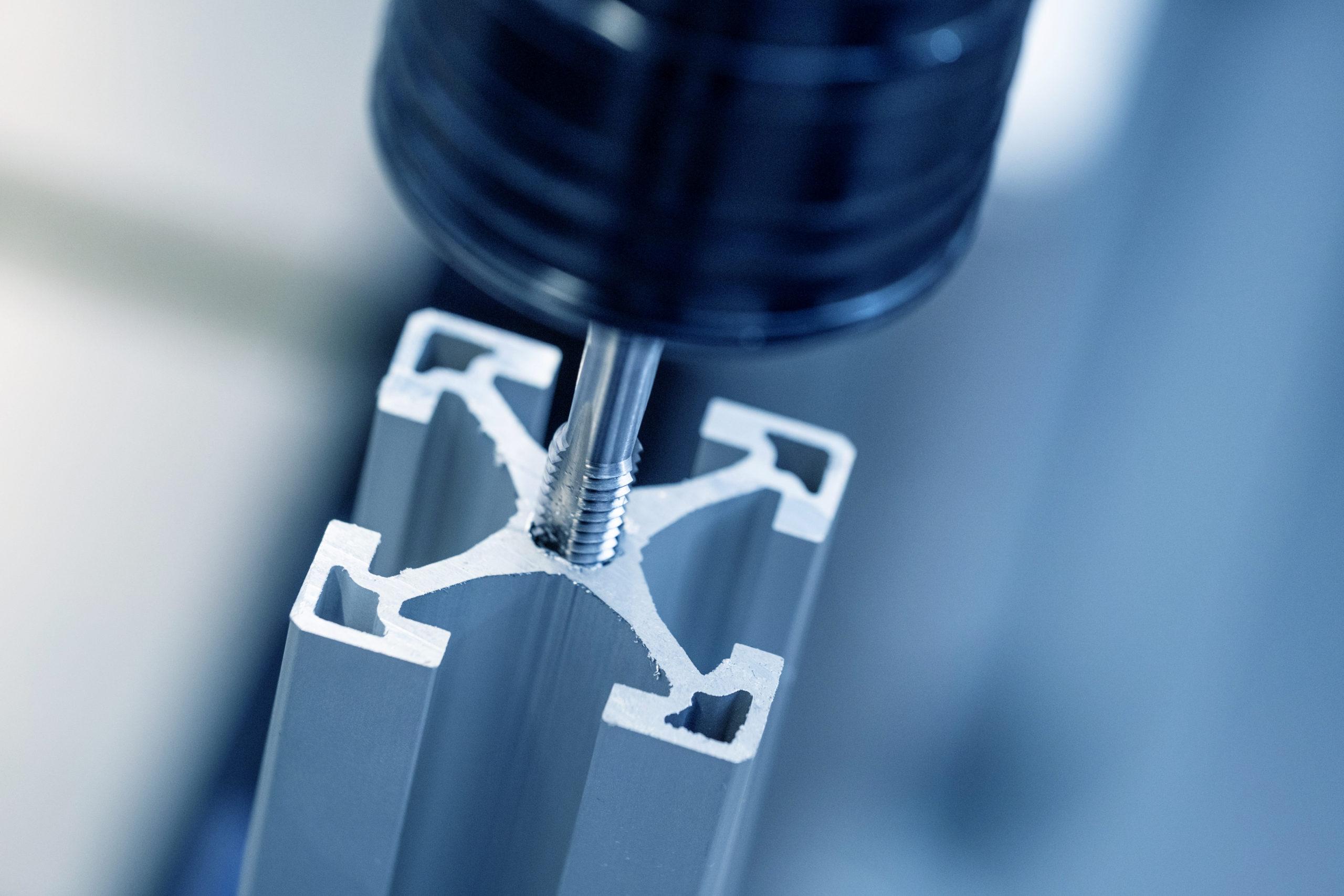 Nahaufnahme einer CNC-Maschine, die ein Gewindeloch in ein Aluminium-Strangpressprofil mit X-förmigem Querschnitt bohrt.