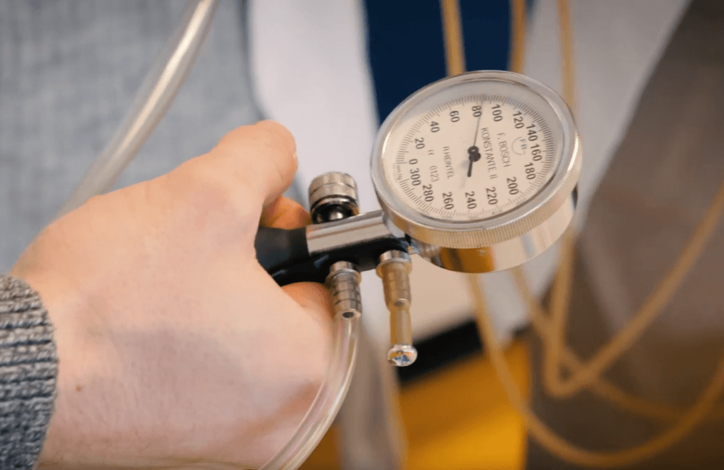 Eine Person hält ein Sphygmomanometer zur Blutdruckmessung in der Hand, an dem Schläuche befestigt und Zahlen auf der Skala sichtbar sind.
