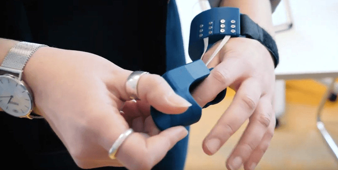 Nahaufnahme einer Person, die ein blaues Armband oder eine Smartwatch anlegt. Die Person trägt silberne Ringe und eine silberne Uhr an ihrer anderen Hand. Der Fokus liegt auf ihren Händen und dem Armband.